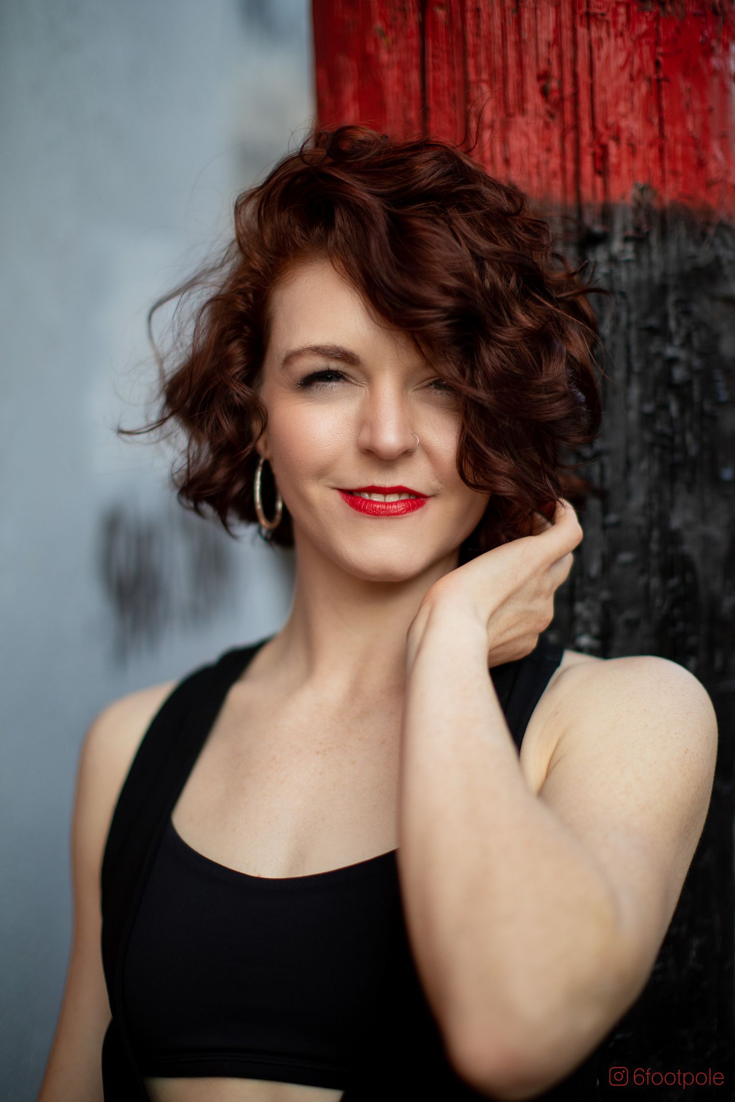Portrait against red and black wall showing curly brown hair, black top, and bold red lipstick.