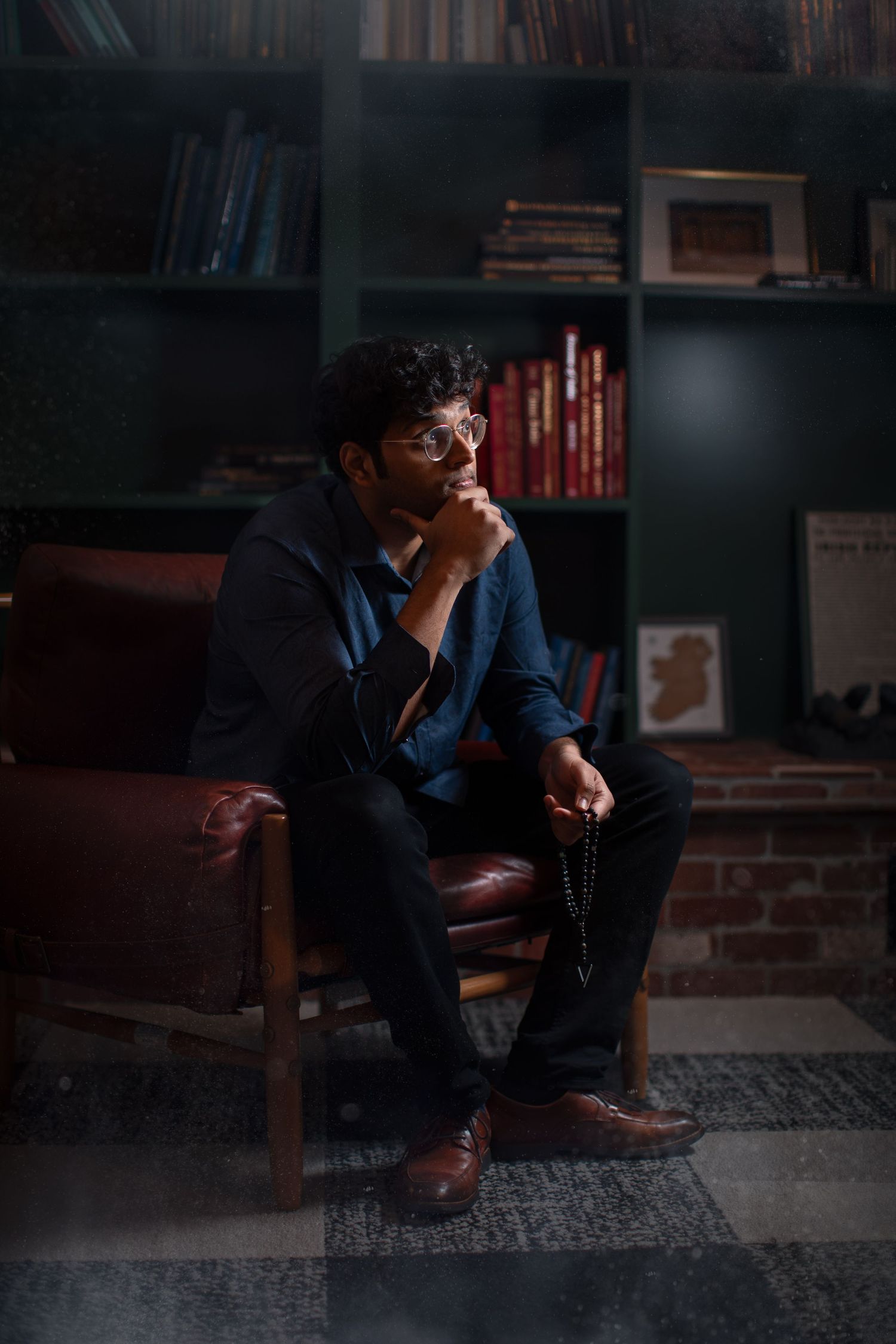 Person wearing glasses sits thoughtfully in a dimly lit library setting with bookshelves in the background.