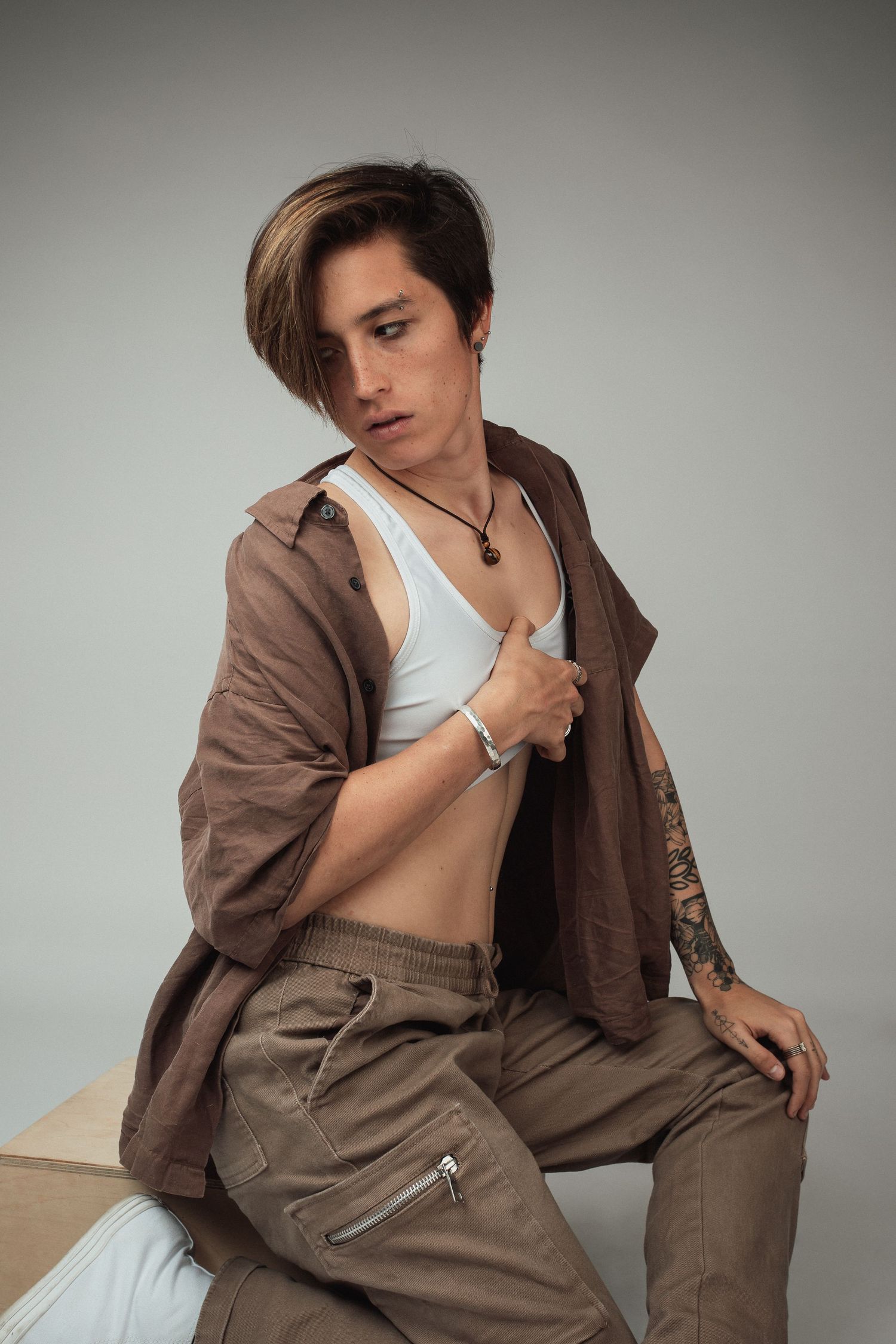 Person in brown jacket and white tank top poses against neutral background in studio.