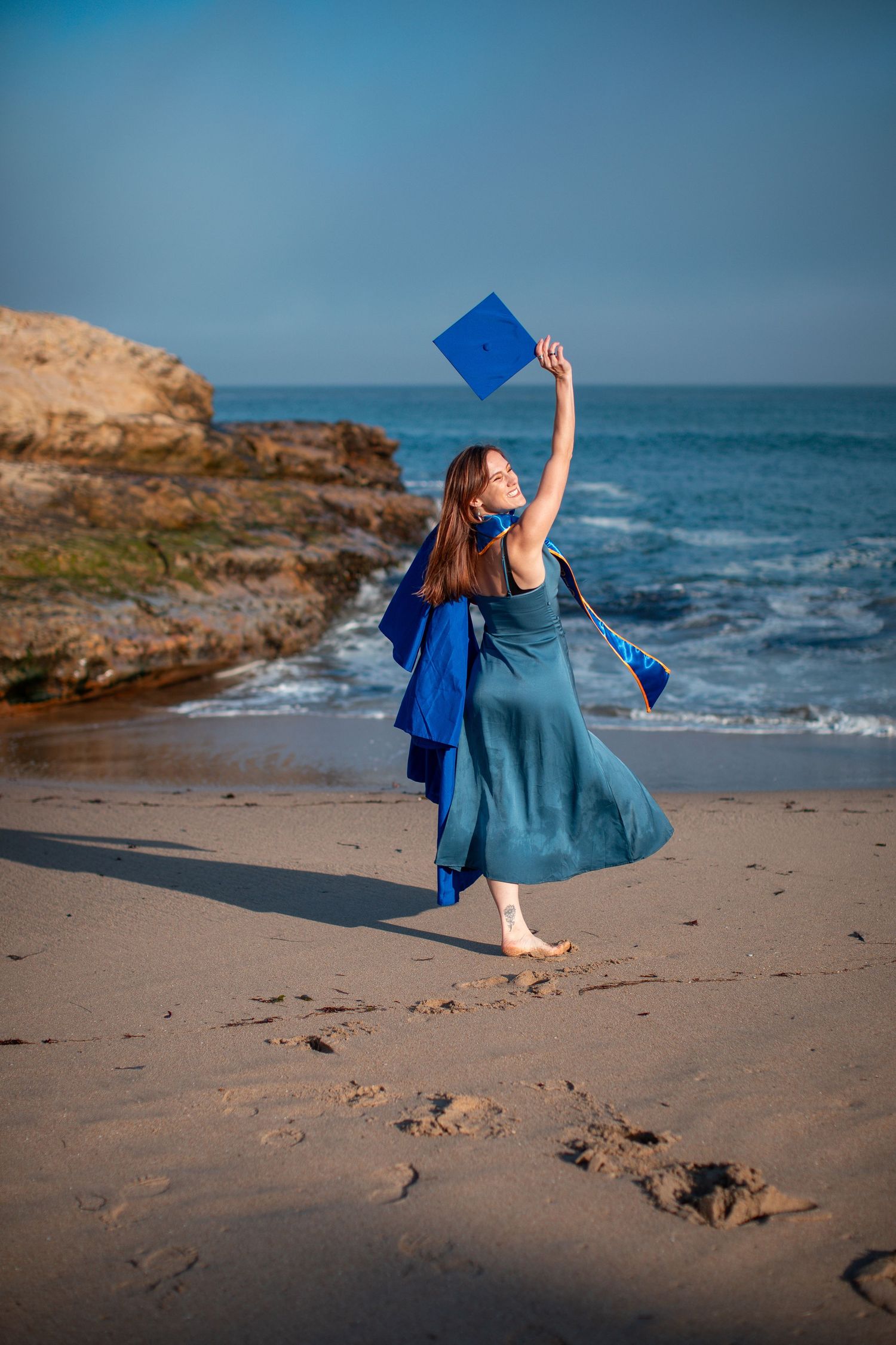 Graduate in teal dress celebrates on beach holding cap against ocean backdrop.
