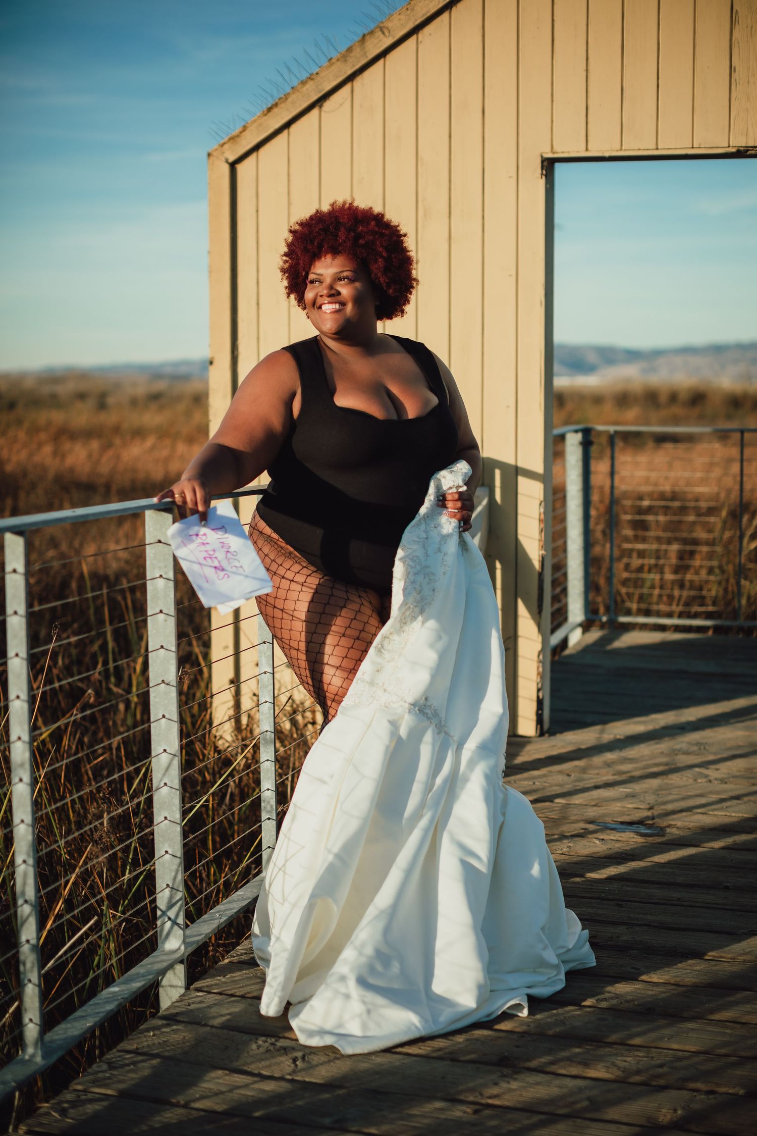 A person in a black top poses with a white dress on a wooden deck during golden hour.