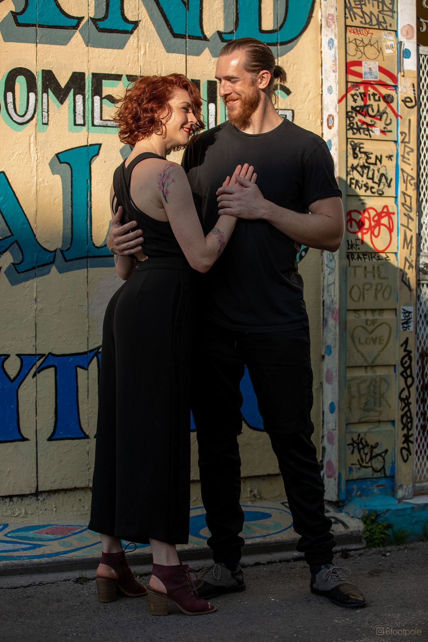 Dancing couple performs against graffiti wall in urban setting wearing black outfits.
