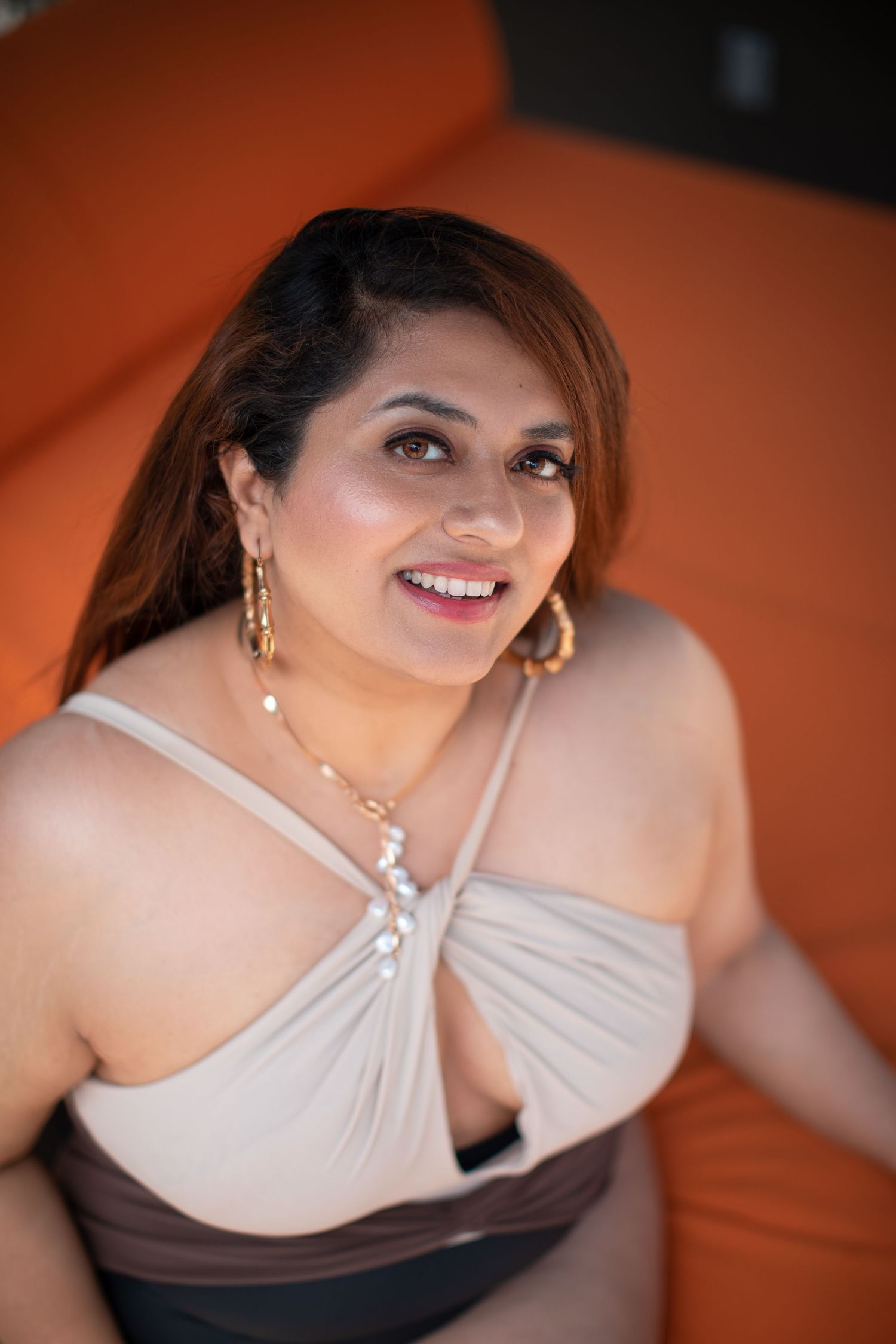 Elegant portrait featuring gold jewelry and a beige halter swimsuit against orange background.