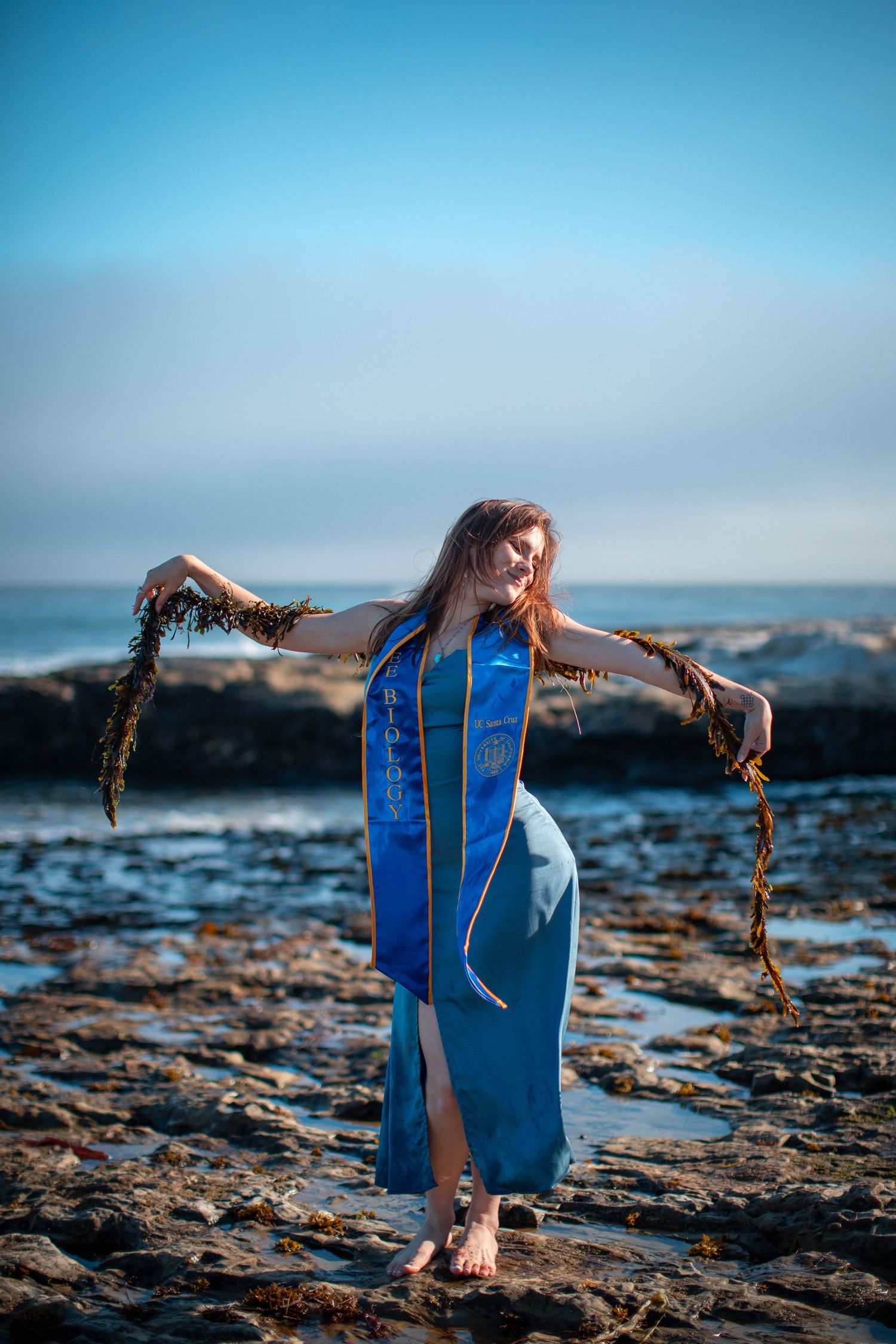 Graduate in blue dress plays with seaweed on rocky beach during sunset photoshoot.