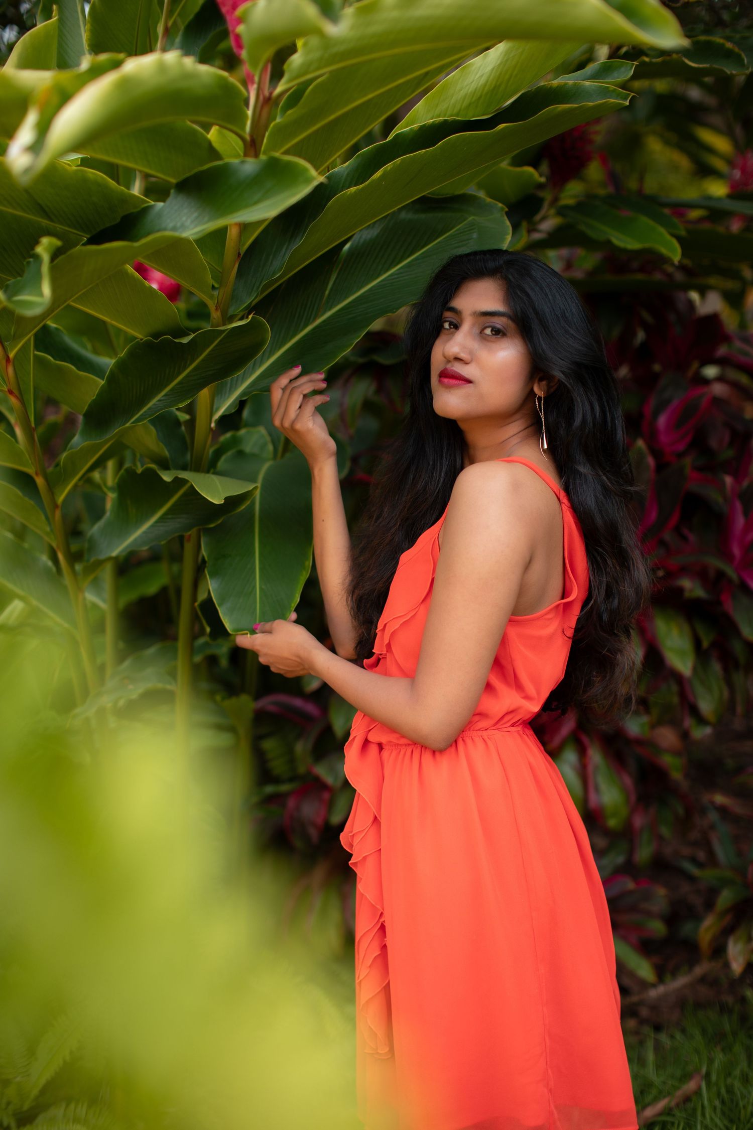 Fashion portrait featuring bright coral dress amidst green tropical garden setting.