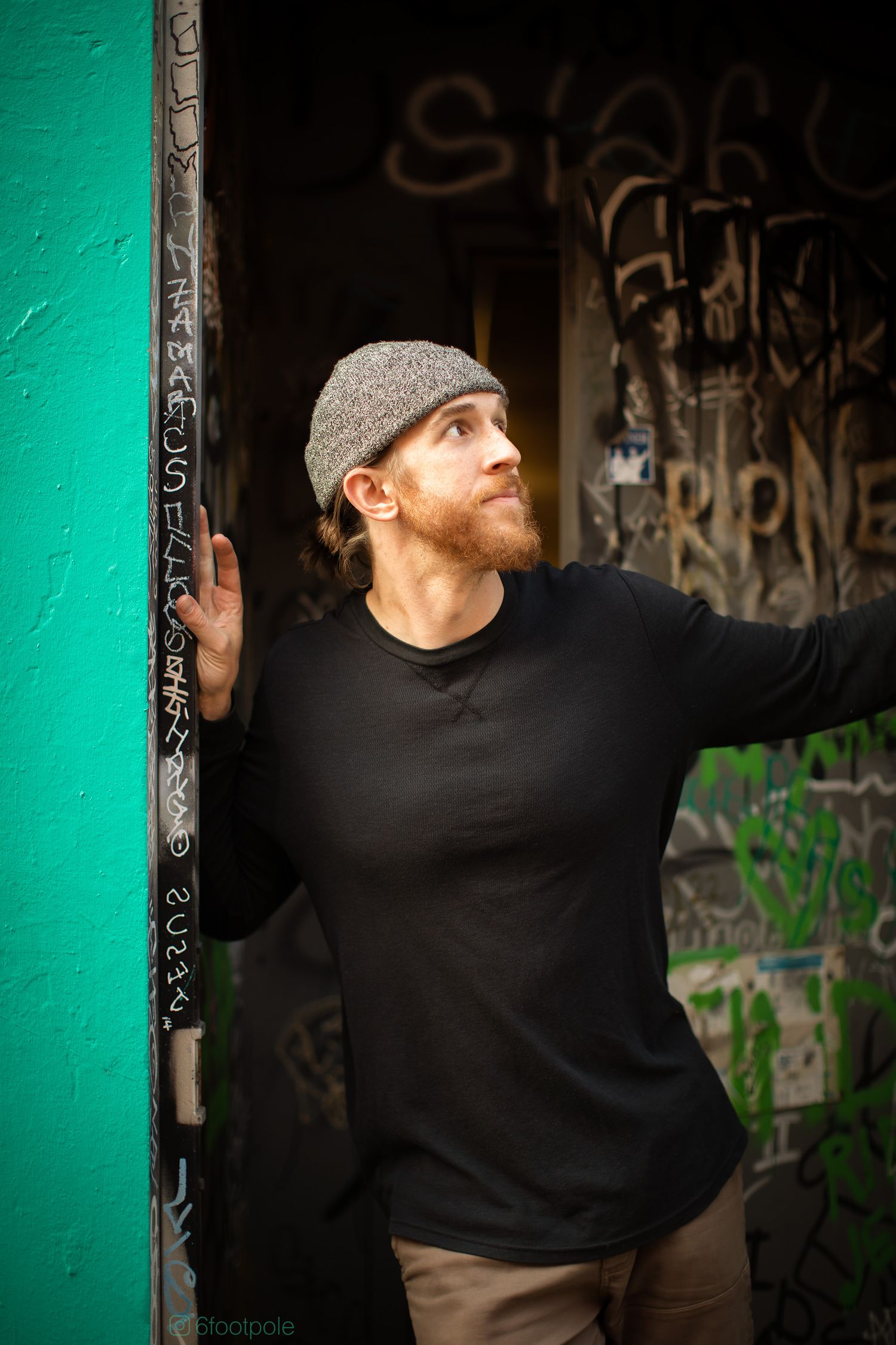 Person in black sweater and gray cap poses against turquoise wall with graffiti background.
