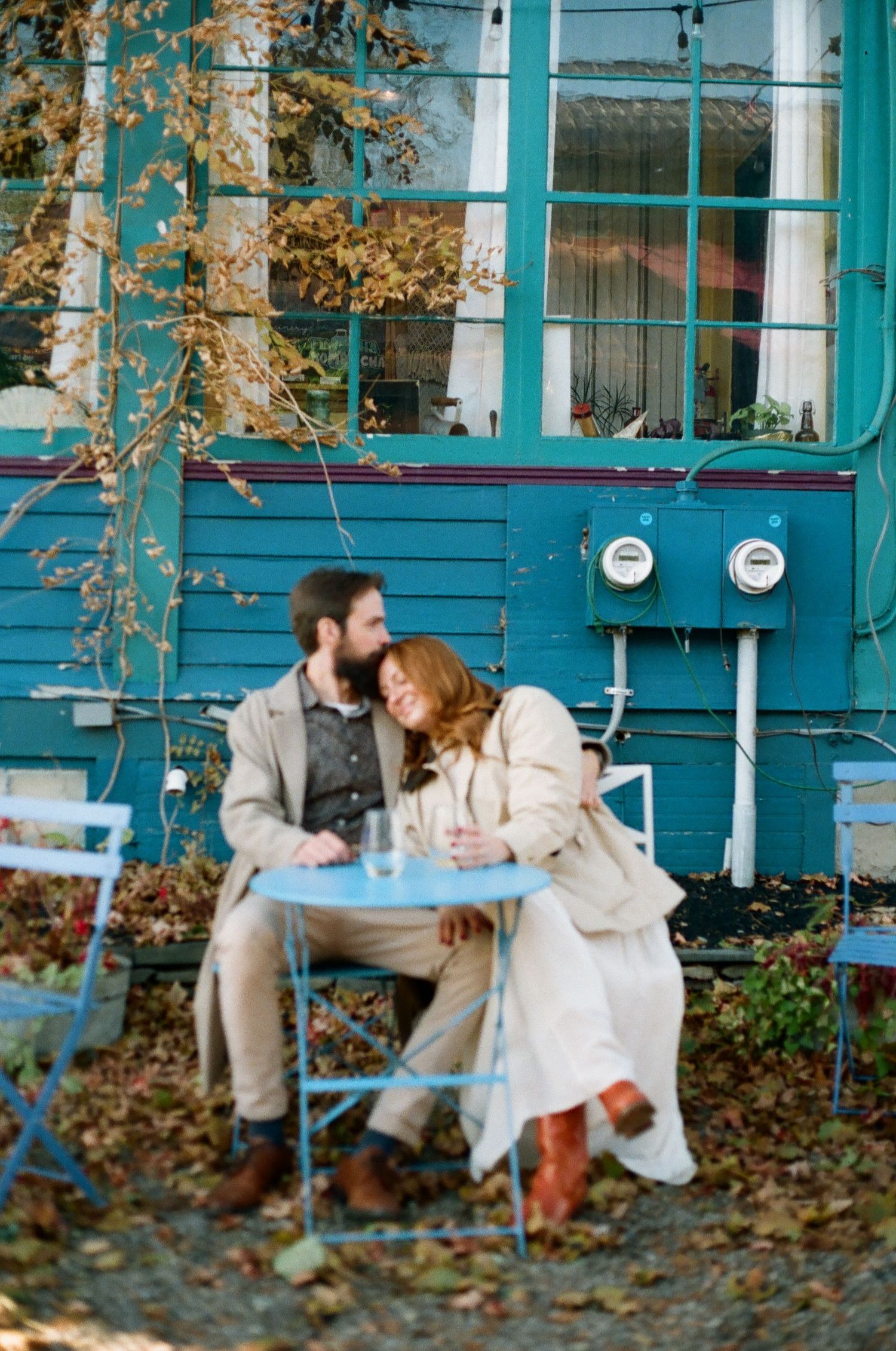 Couple sitting close together at a blue cafe table outside a teal building surrounded by autumn leaves.