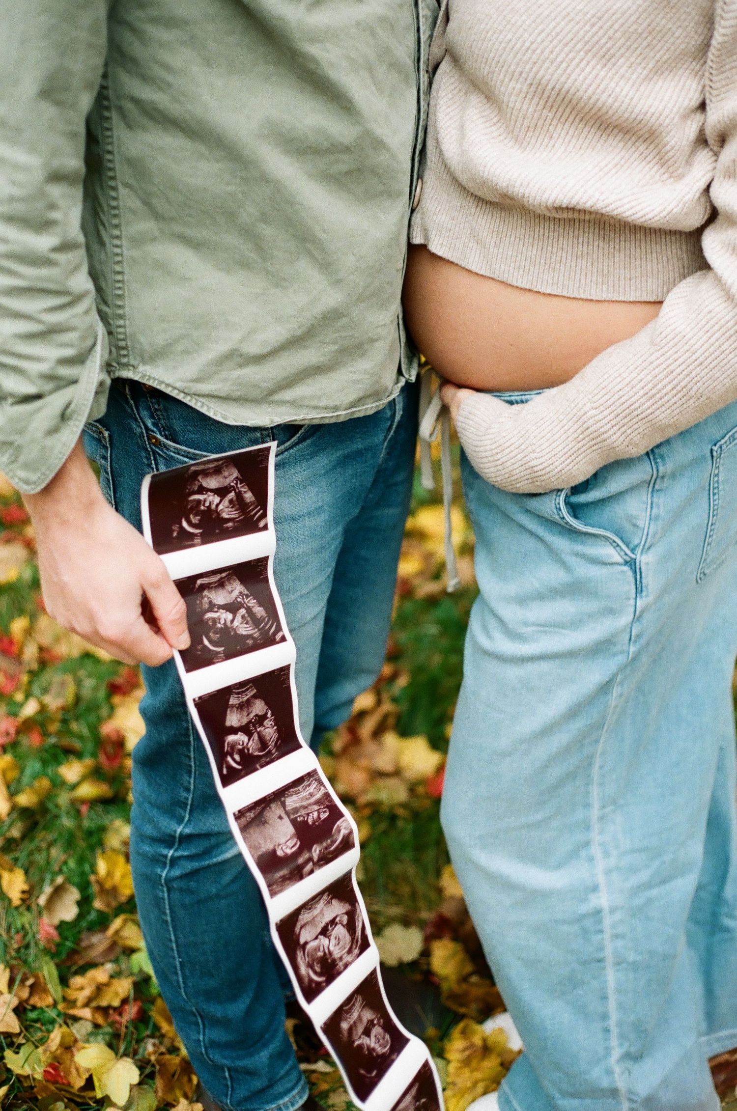 Pregnant couple holding ultrasound strip photos outdoors in autumn, woman's baby bump visible, fall leaves on ground.