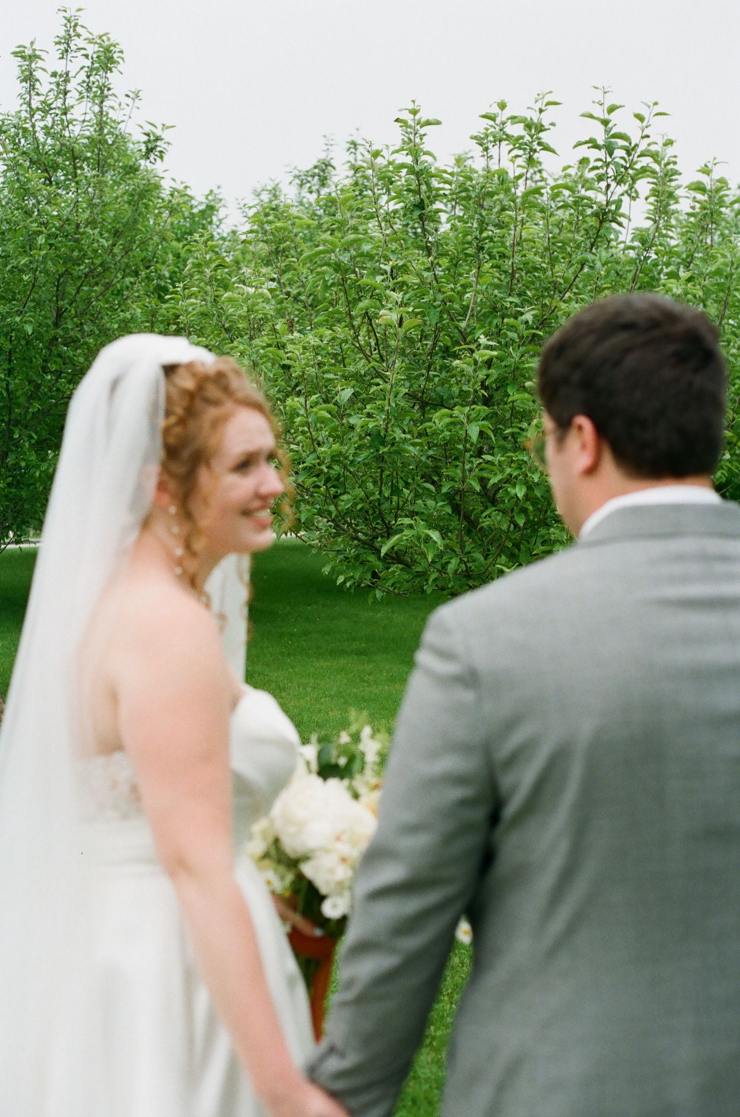 Bride in white gown and veil smiles at groom in gray suit outdoors with lush green trees in background.