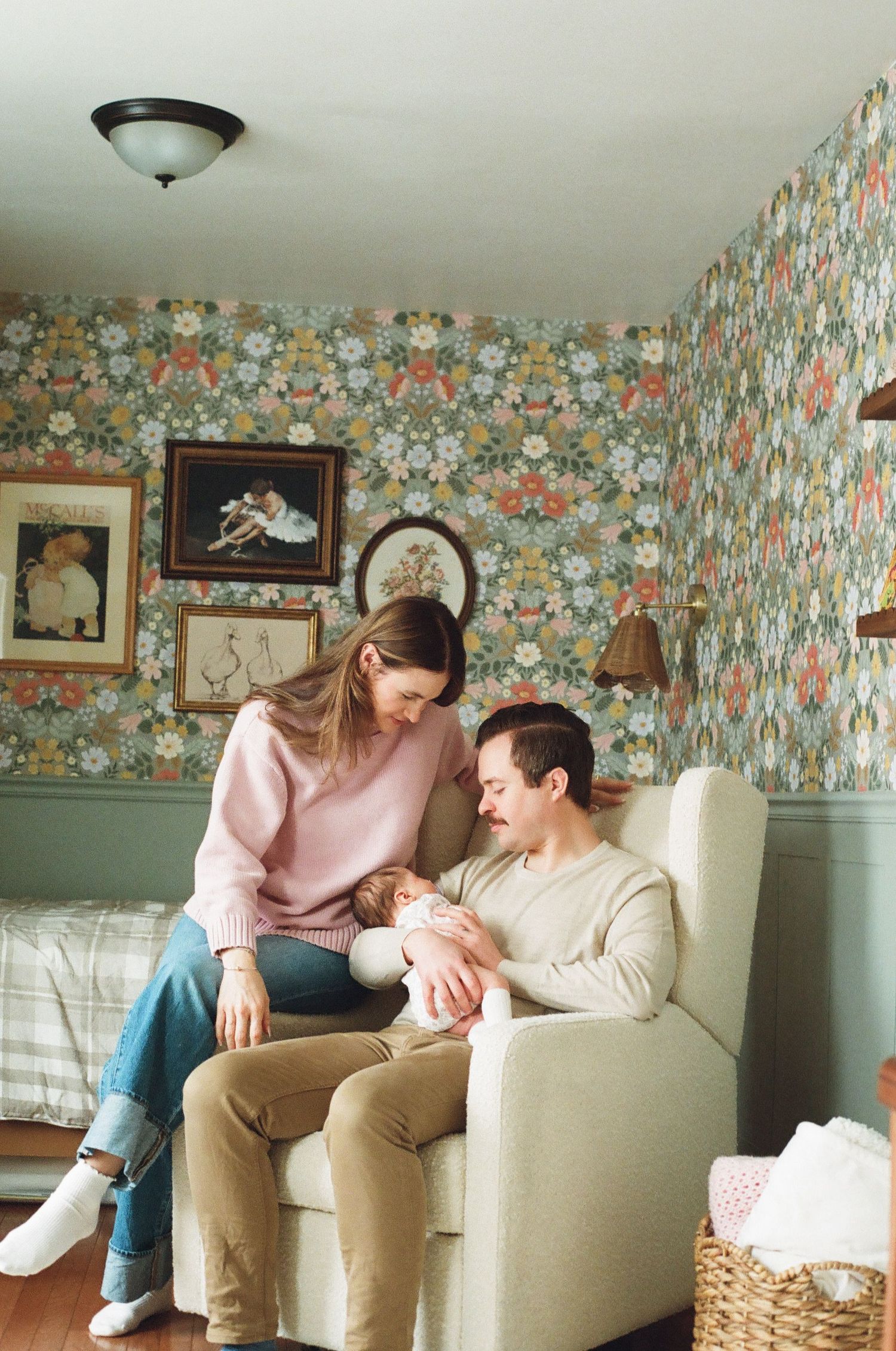 New parents sit in a nursery chair with their newborn, surrounded by floral wallpaper and vintage artwork.