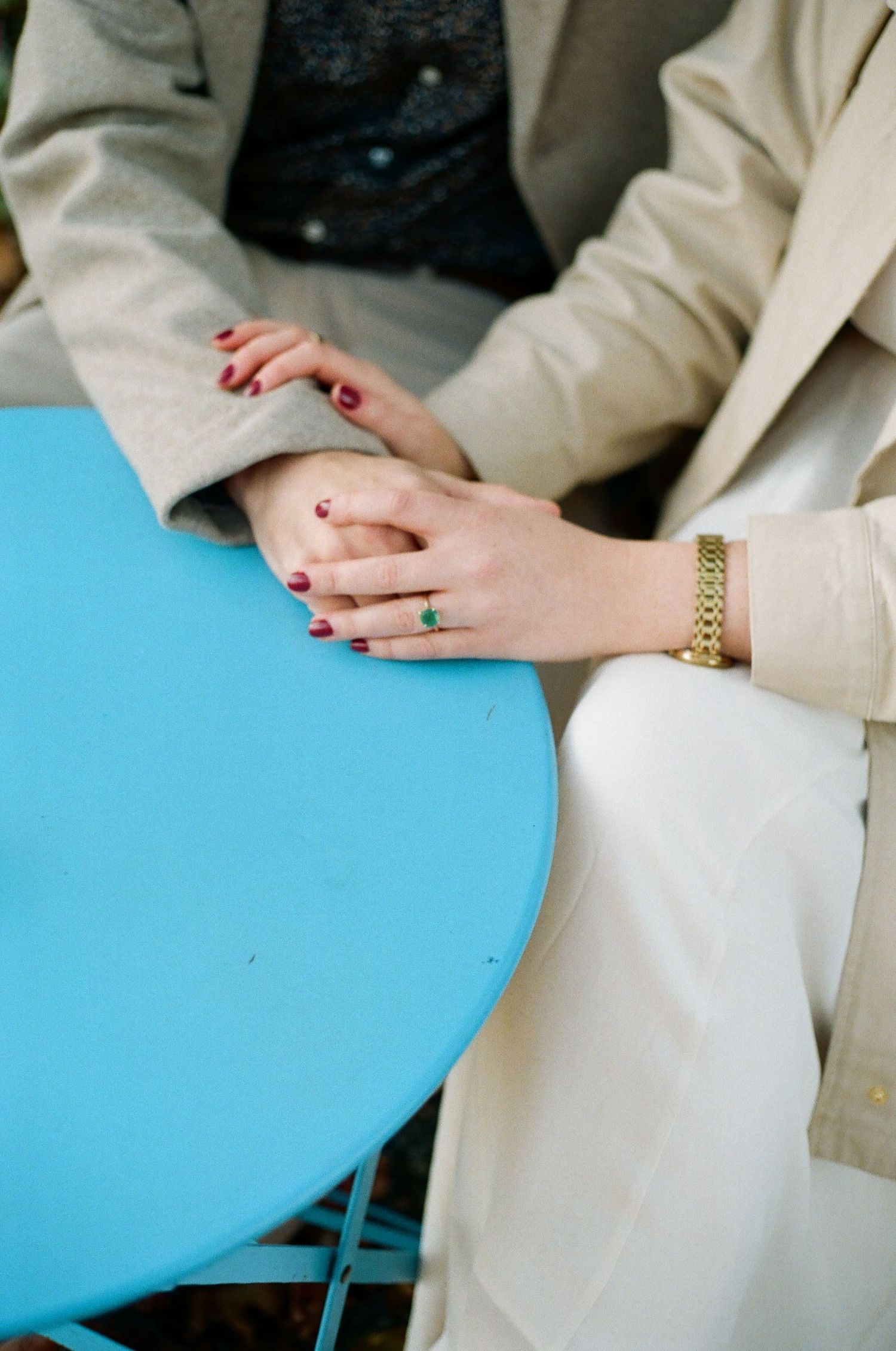 Two people holding hands at a blue table, wearing gold jewelry, emerald ring, and red nail polish.