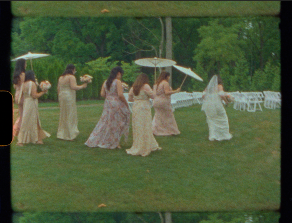 Bridesmaids in pastel gowns gather on a green lawn with parasols at an outdoor wedding ceremony.