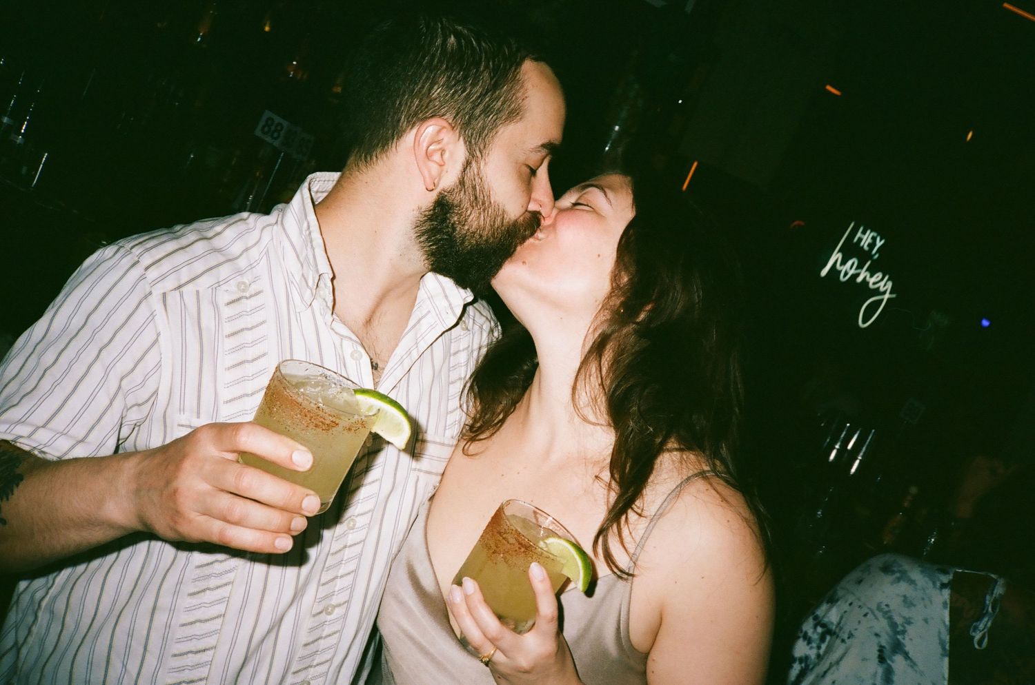 Couple sharing a romantic kiss at a bar while holding cocktails with lime garnishes.