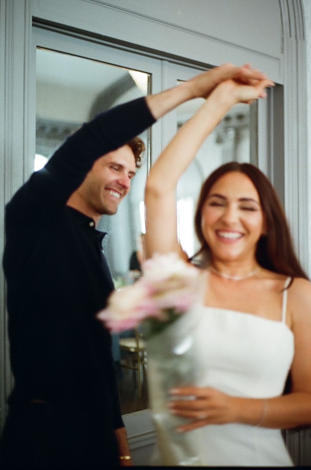 A joyful couple dancing together indoors, man in black shirt spinning woman in white dress holding pink flowers.