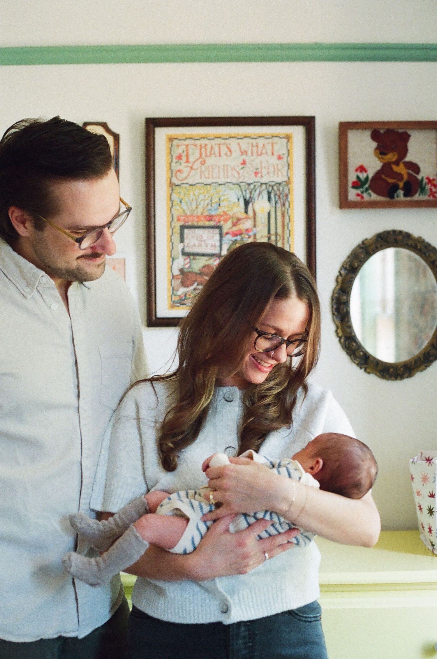 New parents in glasses admire their newborn baby in a cozy nursery with vintage wall art.