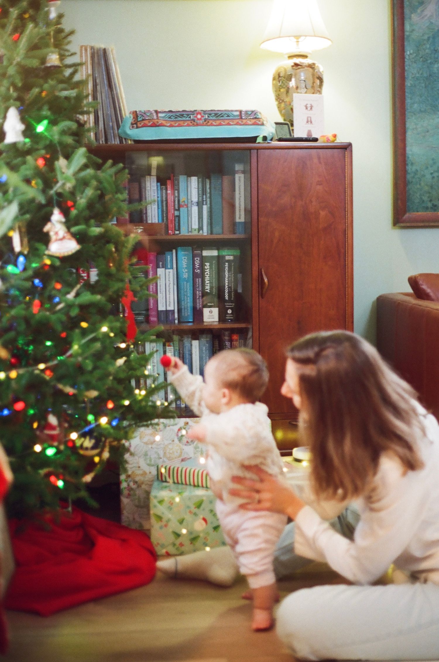 Mother and baby exploring a decorated Christmas tree with colorful lights and gifts beside a wooden bookshelf.
