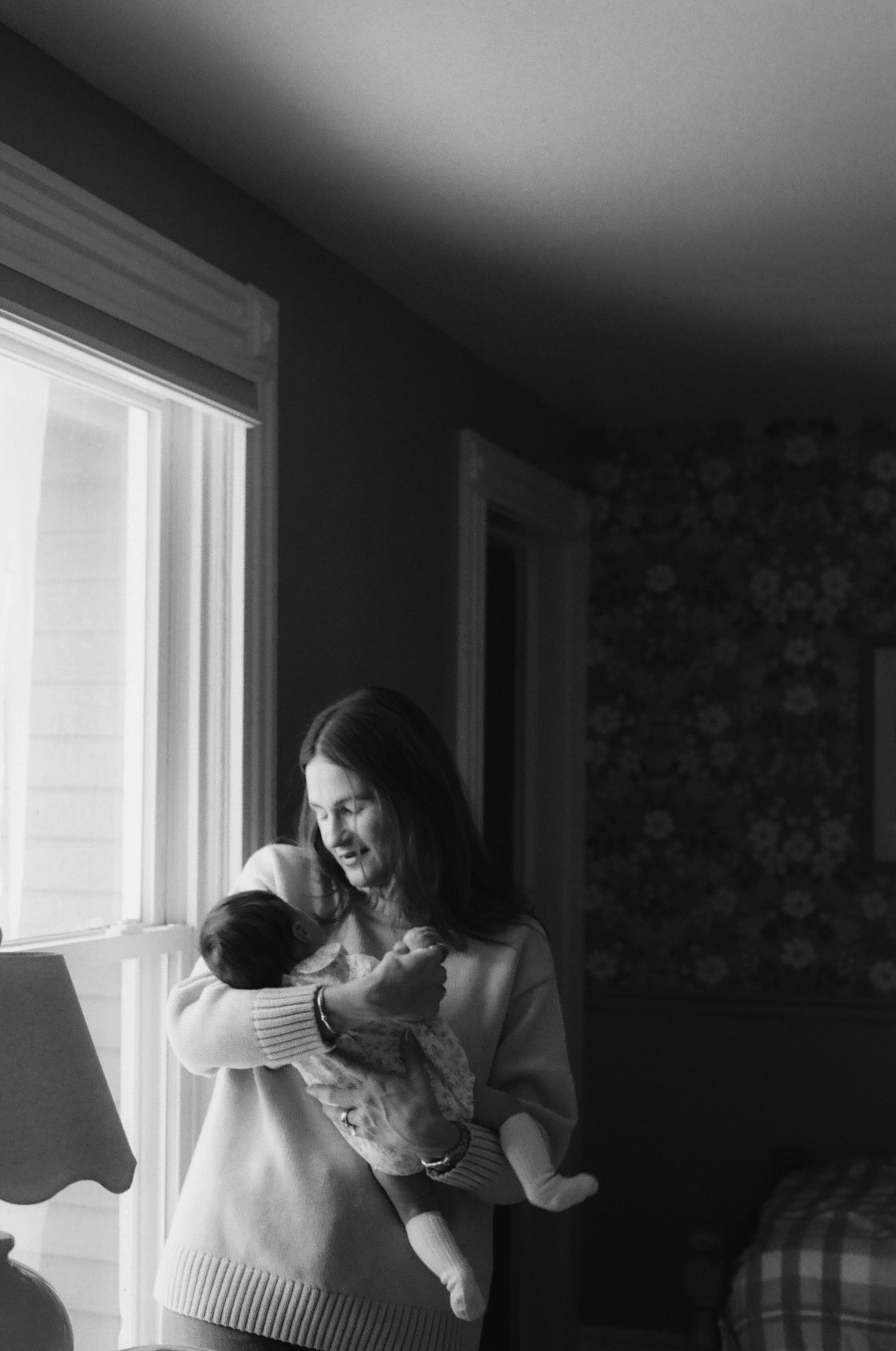 Black and white photo of a smiling woman holding a newborn baby near a bright window indoors.