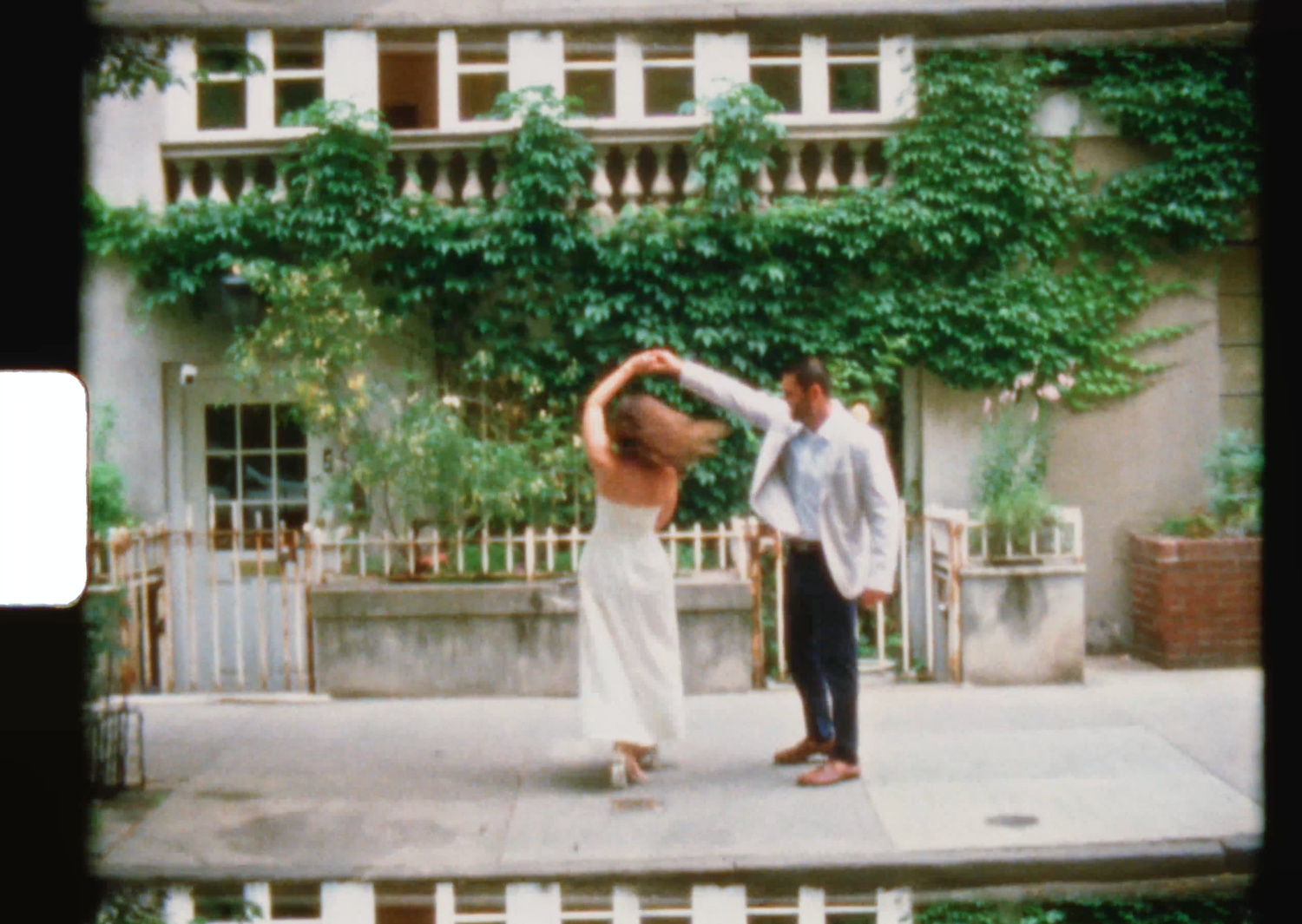 A couple dances outdoors in front of an ivy-covered building, the woman in a white dress twirling gracefully.