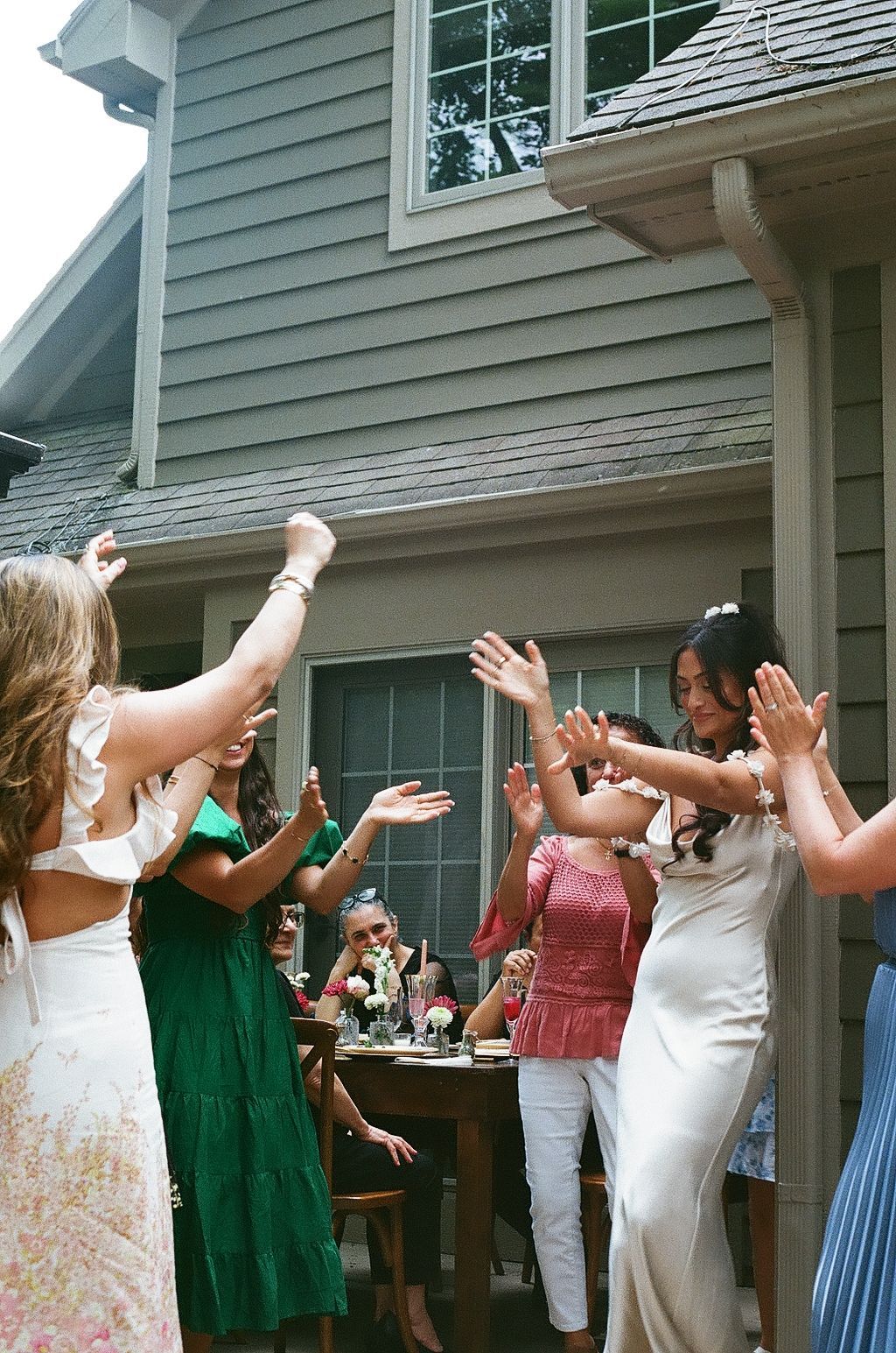 Women in elegant dresses celebrating outdoors at a bridal shower, raising hands joyfully near a decorated table.
