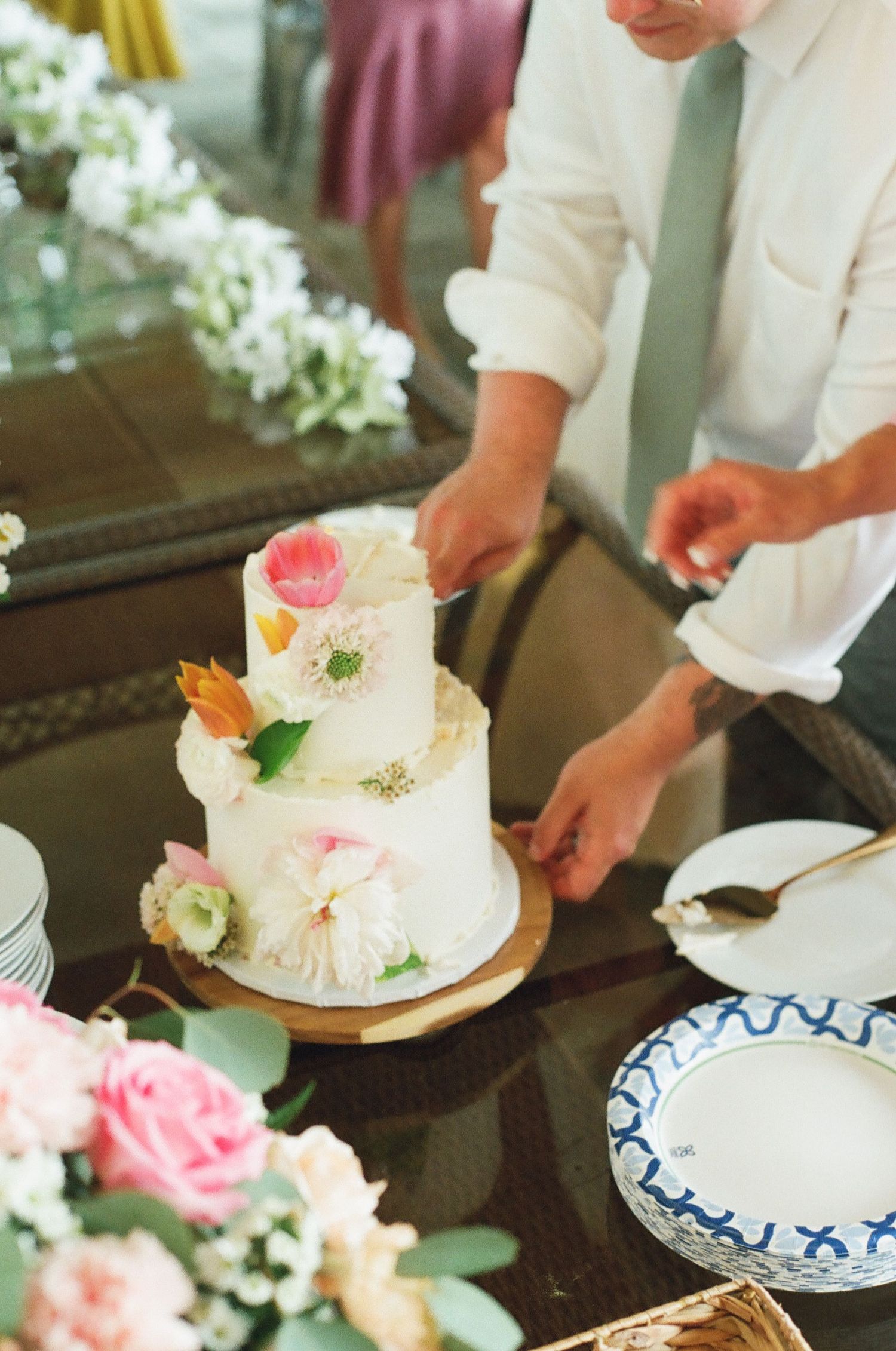Person cutting a two-tier white wedding cake decorated with colorful flowers on a glass table with roses nearby.
