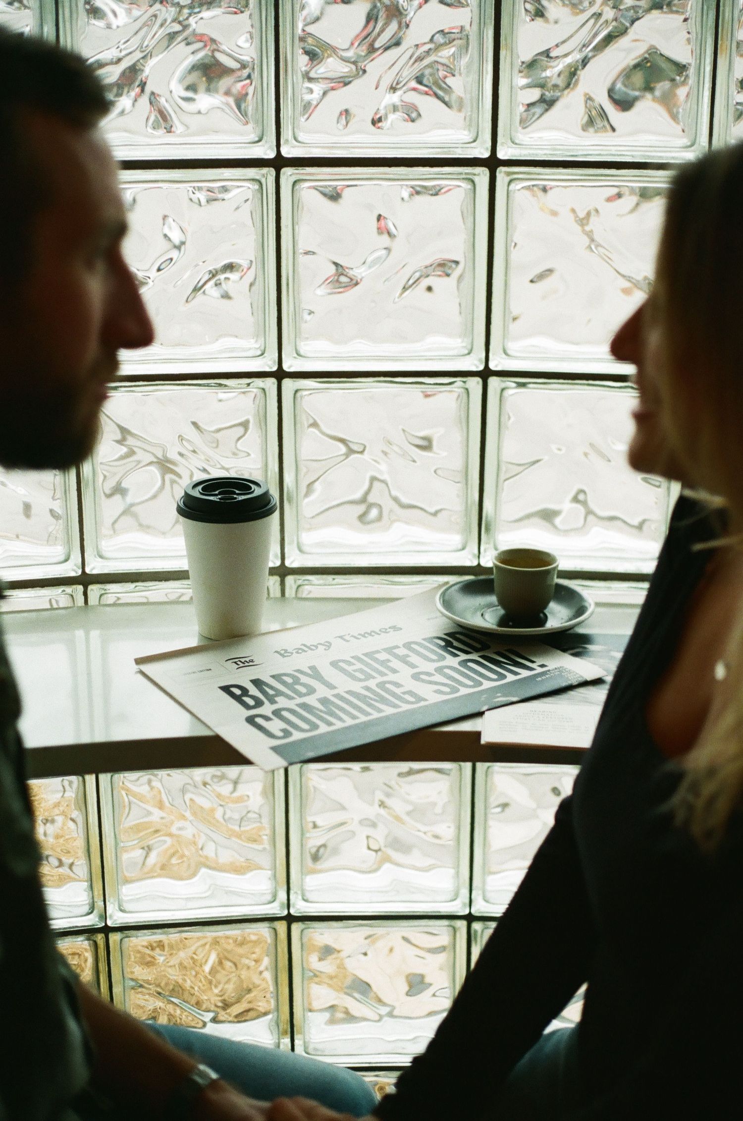 Two people sit facing each other over coffee cups and a Baby Captains Commission newspaper by a glass block wall.
