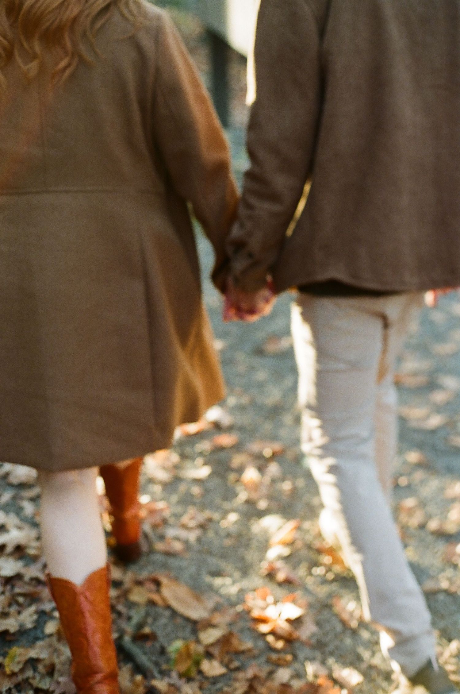 Couple holding hands while walking on a leaf-covered path in autumn, wearing matching brown coats and boots.