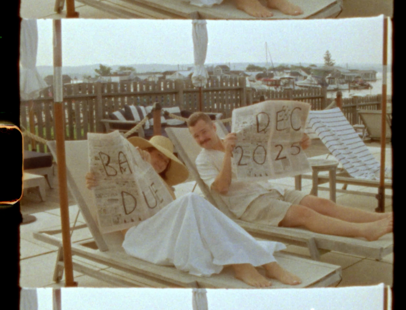 Two people relaxing on beach chairs holding handmade signs reading BAR DU and DEC 2025 in a sunny outdoor setting.