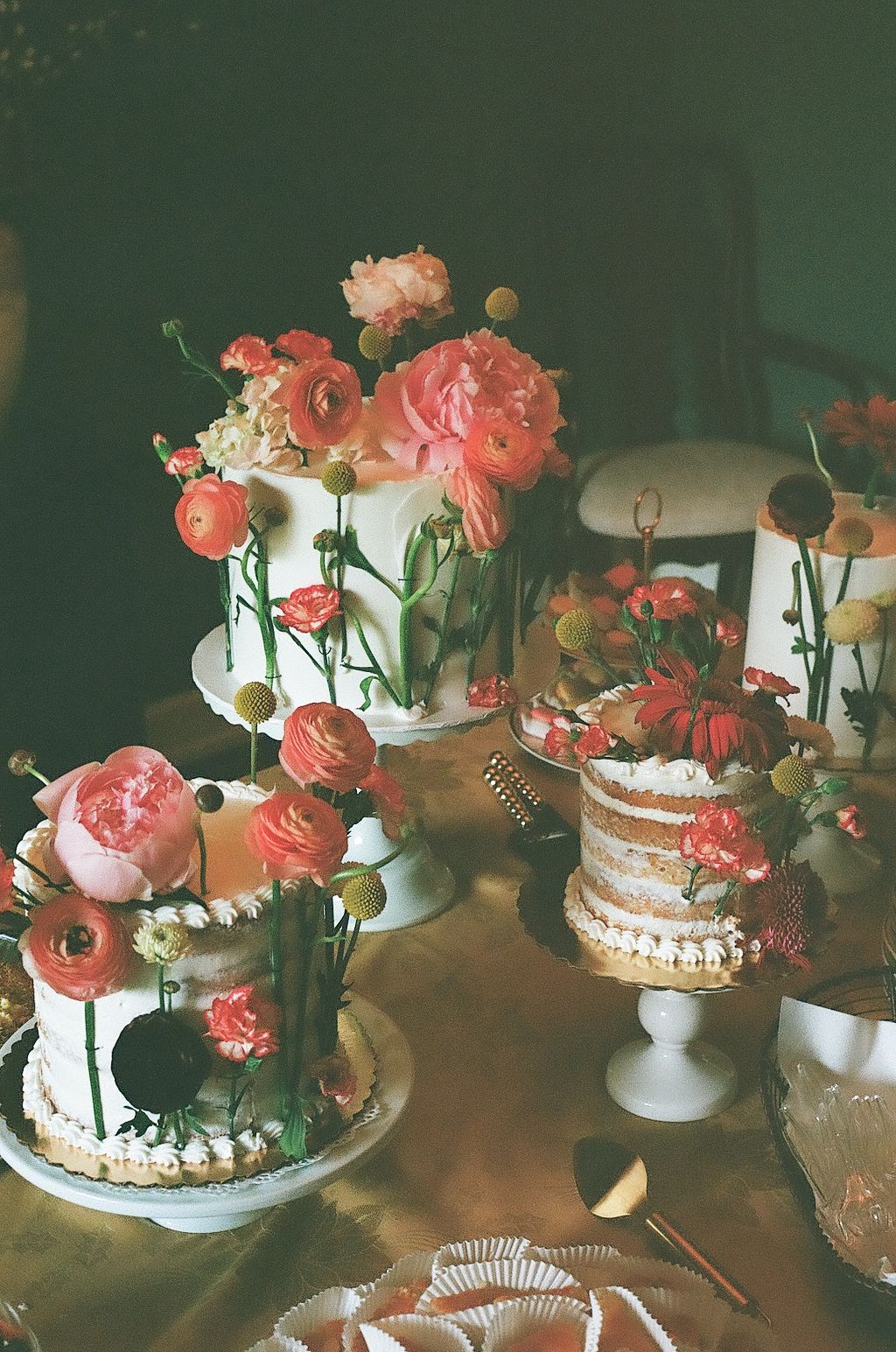 Multiple white wedding cakes adorned with coral and pink ranunculus flowers displayed on pedestals at a rustic dessert table.
