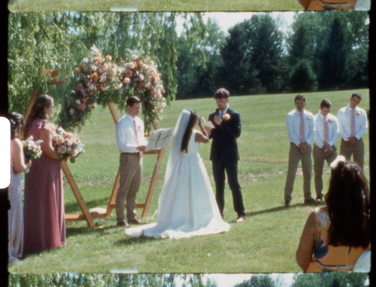 Outdoor wedding ceremony on green lawn with floral arch, bride and groom exchanging vows, wedding party in pink and khaki.