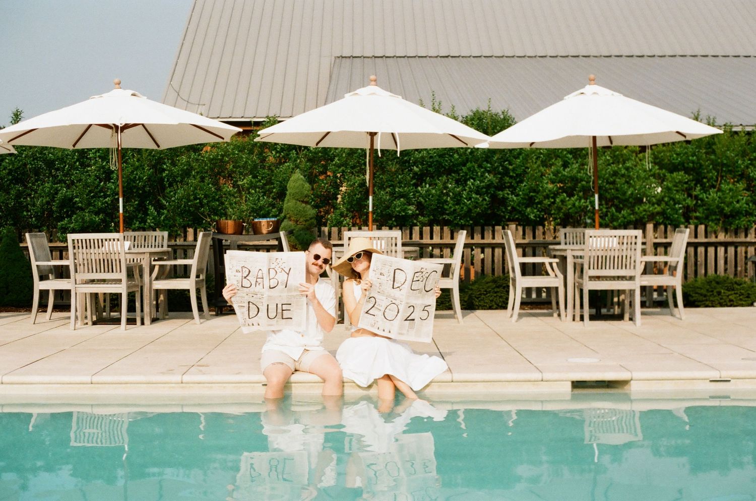 Two women sit poolside holding baby due date signs near white umbrellas and outdoor seating at an elegant venue.