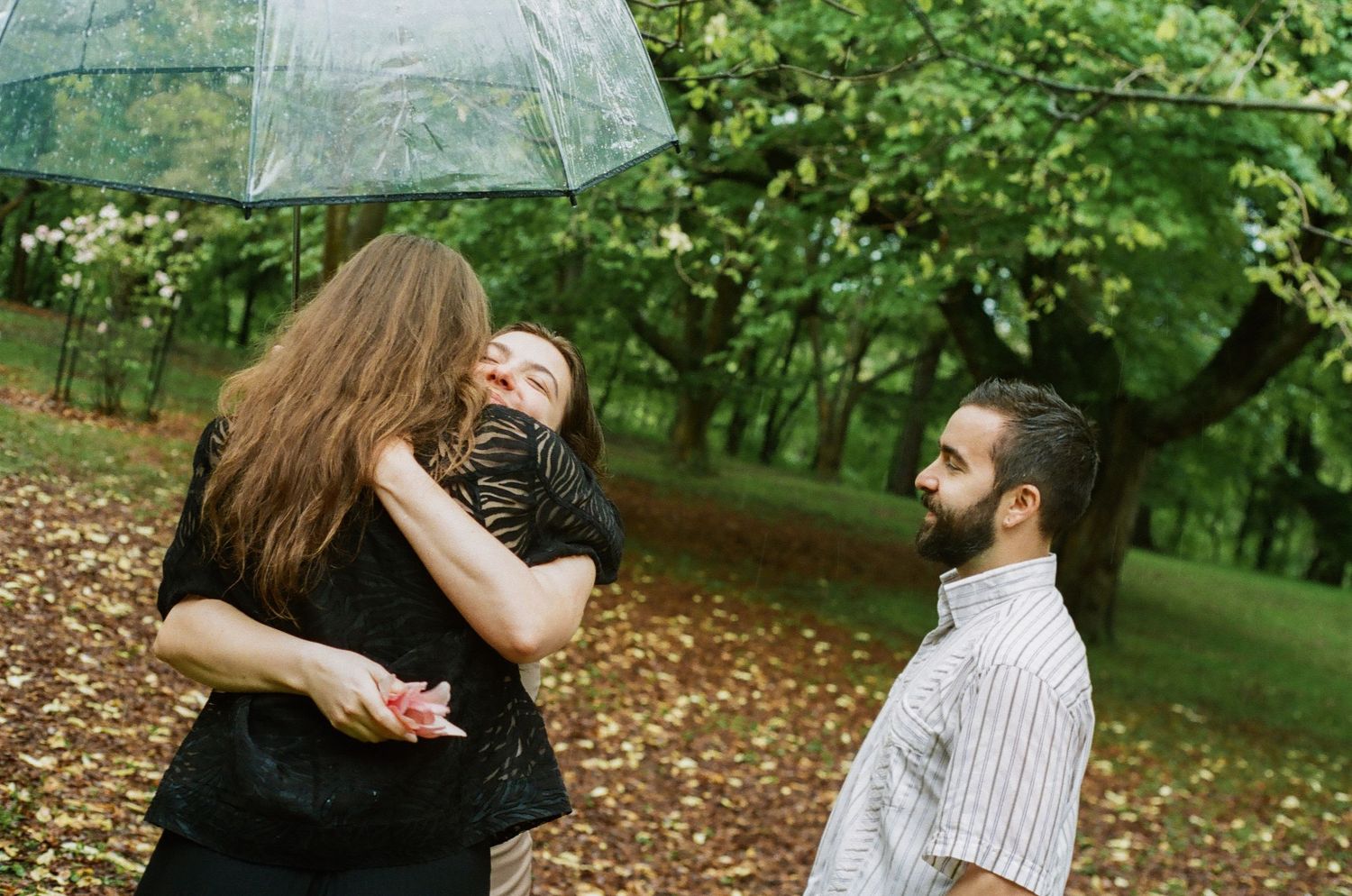 Two women embrace under a clear umbrella in a park while a bearded man watches, surrounded by autumn leaves.