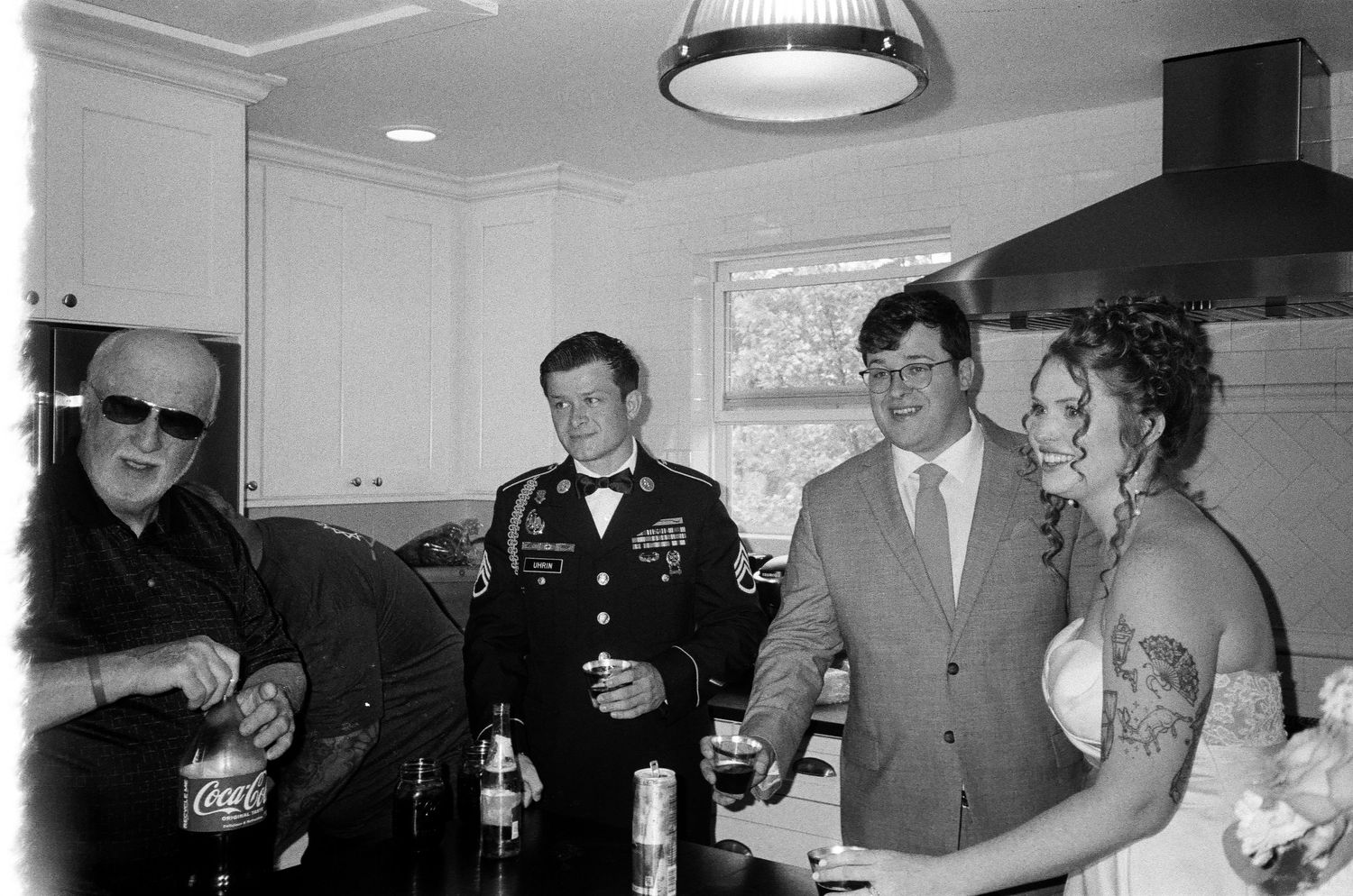 Black and white photo of a bride and groom celebrating with guests, one in military uniform, in a kitchen.