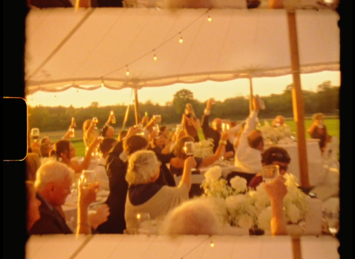 Guests raising glasses in a toast under a white tent at an outdoor wedding reception at golden hour.