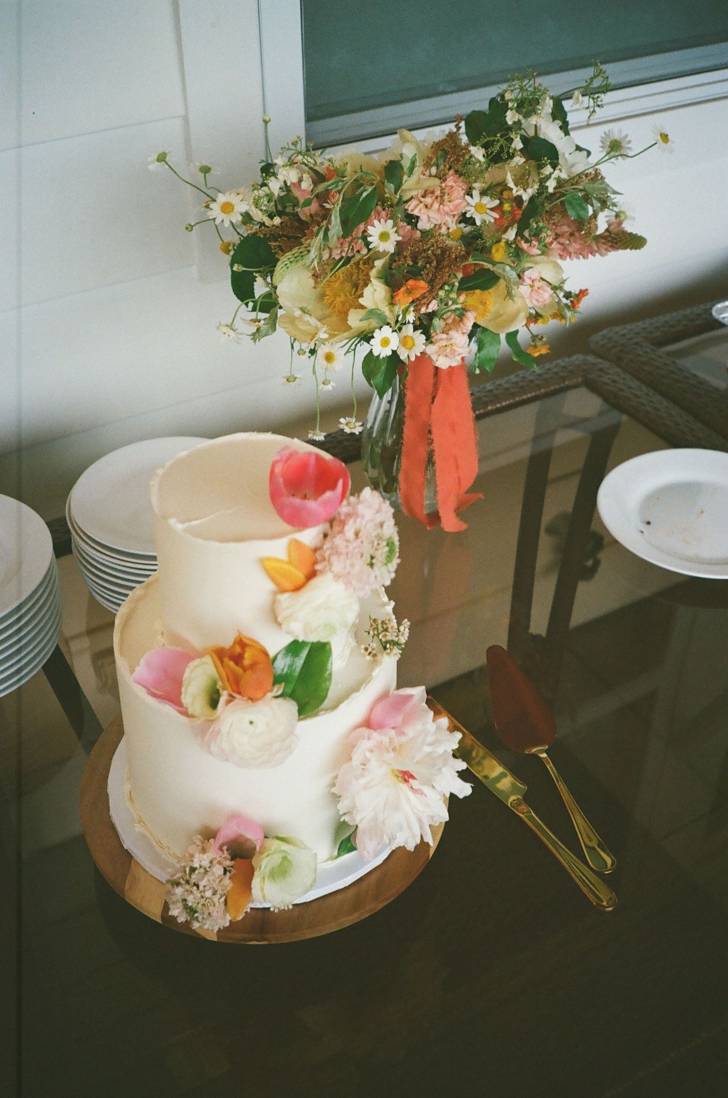Two-tier white wedding cake adorned with fresh colorful flowers on a wooden stand, with a floral bouquet in the background.