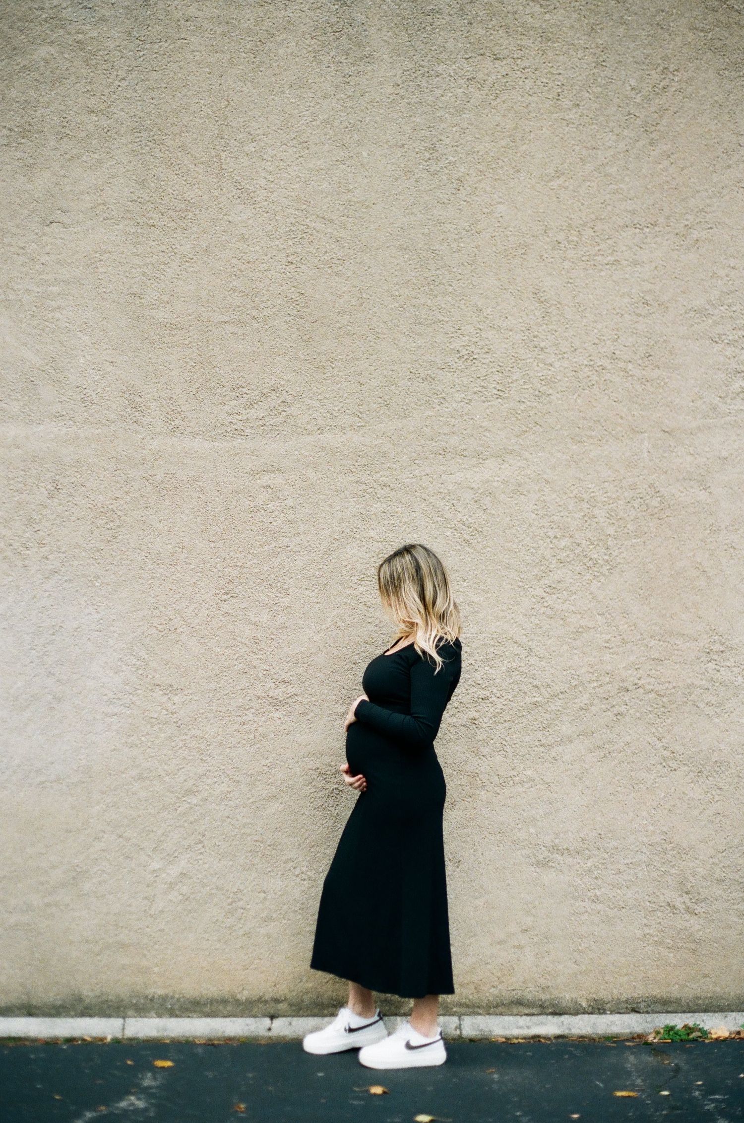 Pregnant woman in black dress and white Nike sneakers standing against a beige stucco wall, cradling her baby bump.