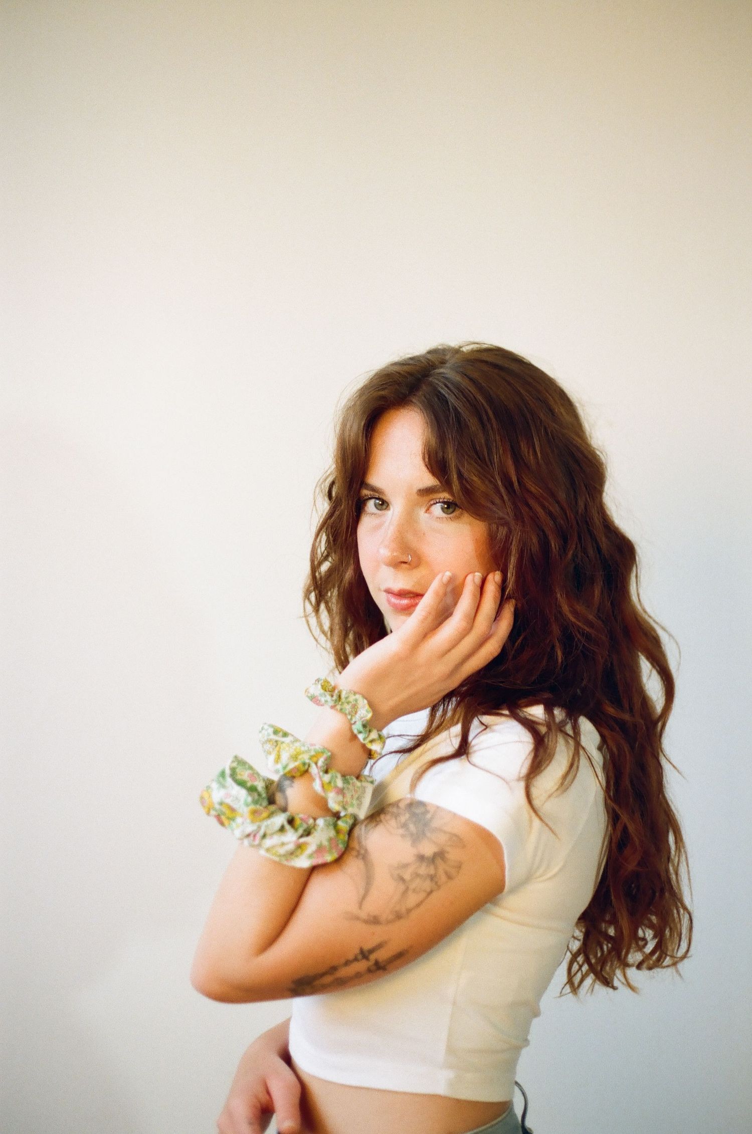 Young woman with curly brown hair in white crop top, floral scrunchie bracelet, and arm tattoo posing against white background.