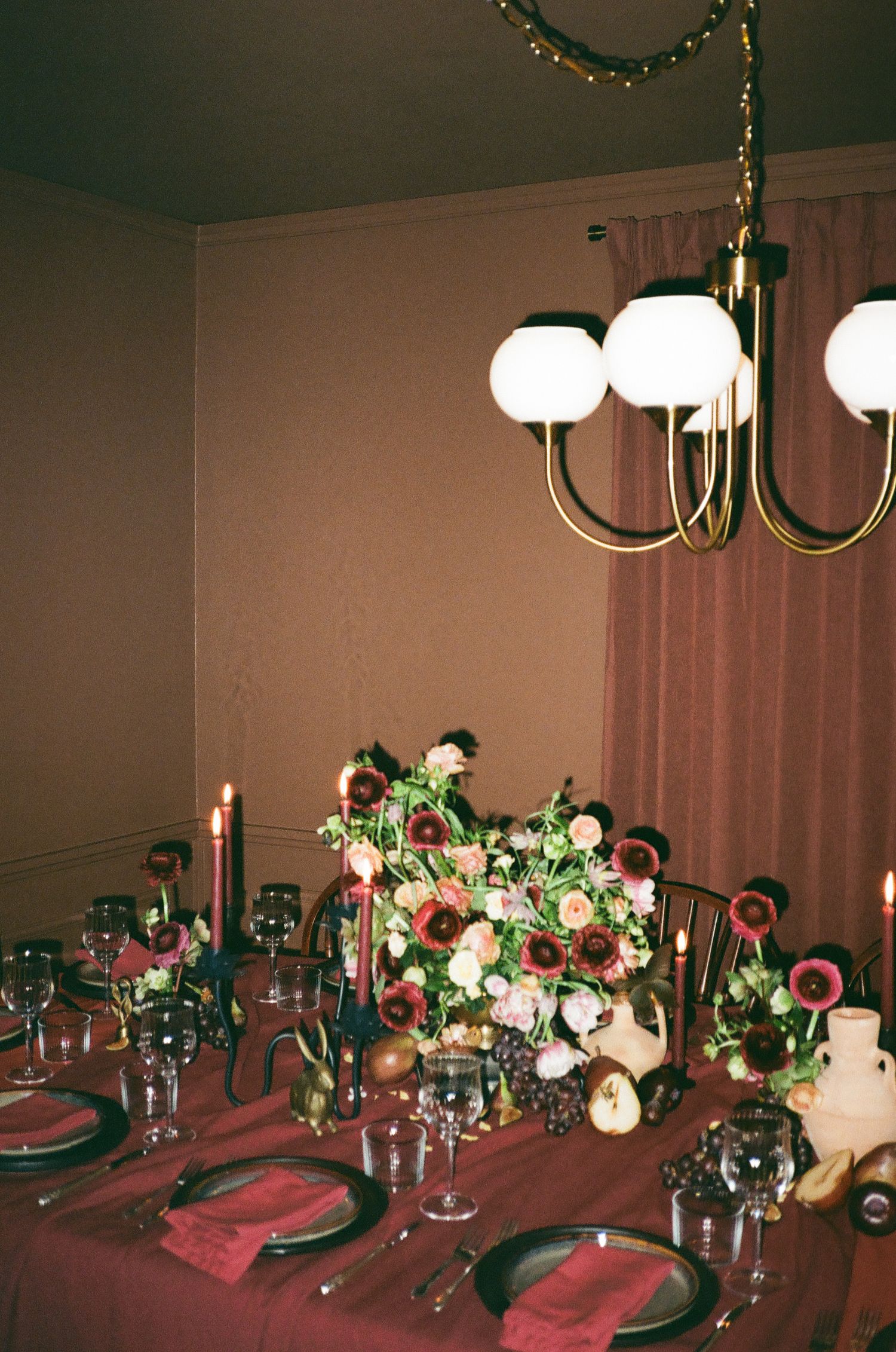 Elegant dining table with floral centerpiece, candles, dark plates, and wine glasses under a gold chandelier.