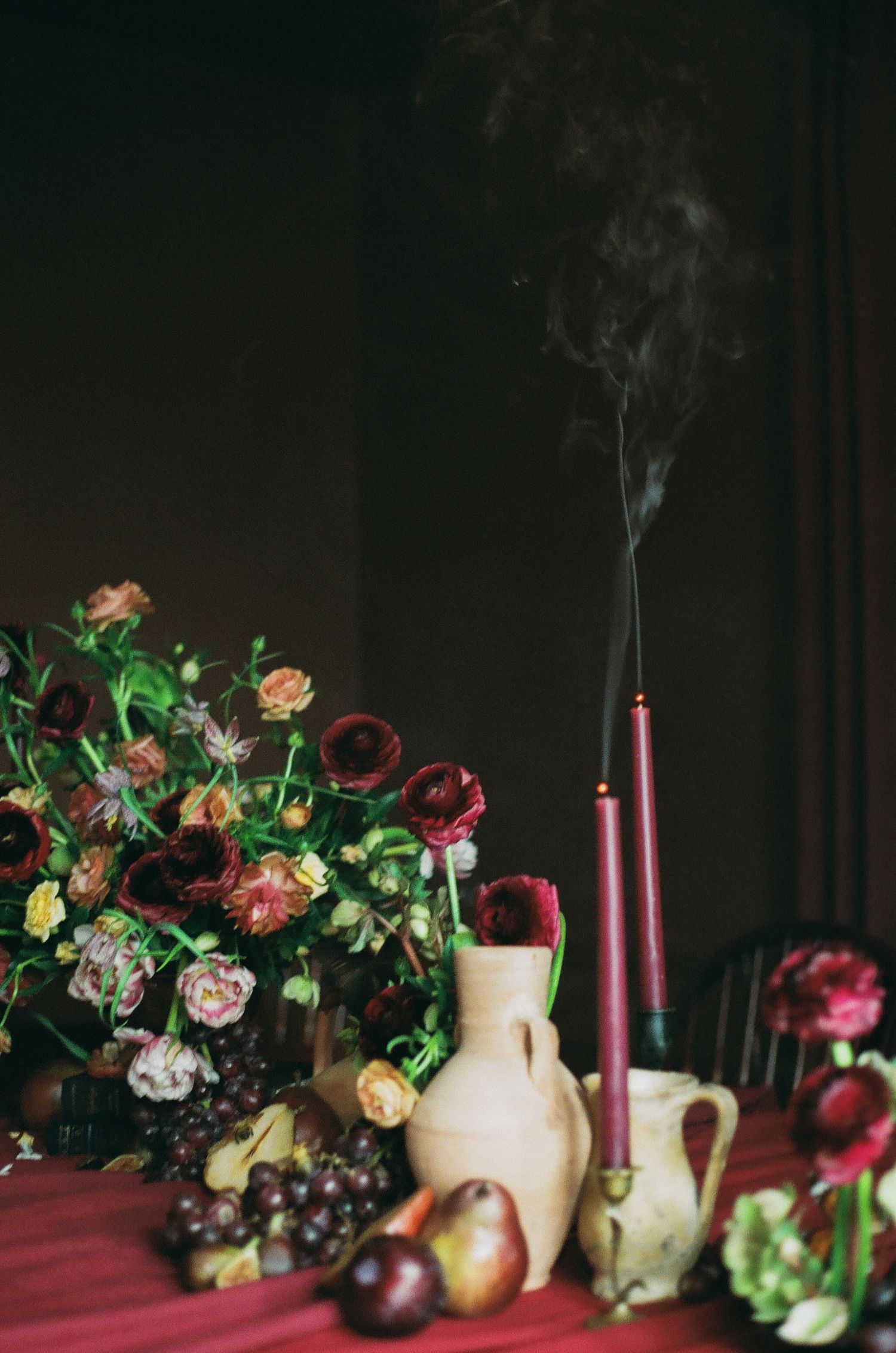 Dark floral arrangement with burgundy candles, ceramic vases, and fresh fruit on a red tablecloth.