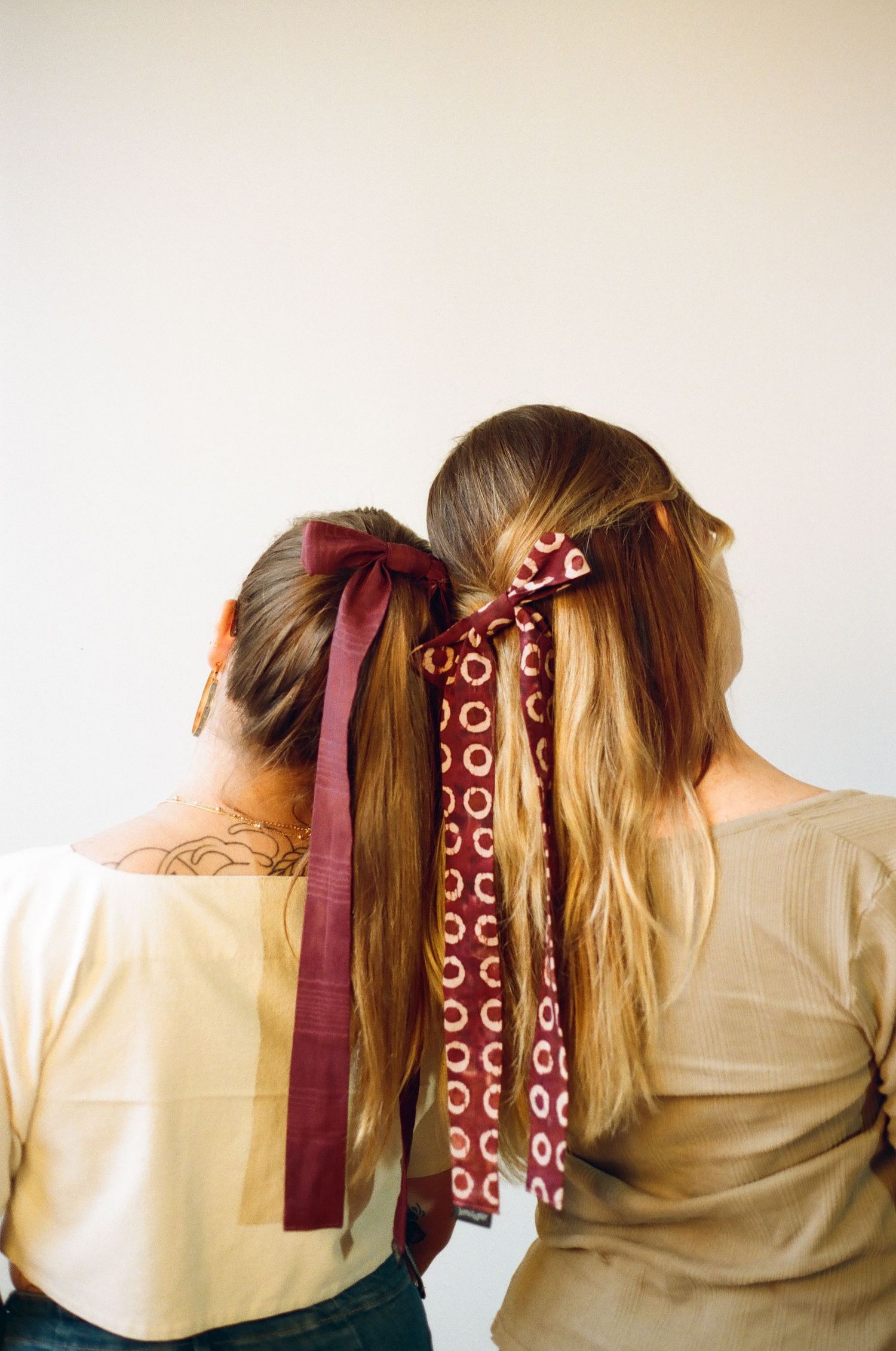 Two women with long hair styled in ponytails adorned with burgundy ribbon bows, viewed from behind.