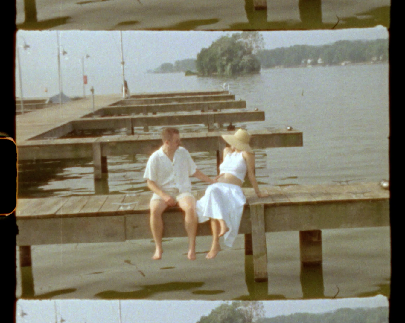 Vintage photo of a couple sitting on a wooden dock by a lake, dressed in white summer clothing on a sunny day.