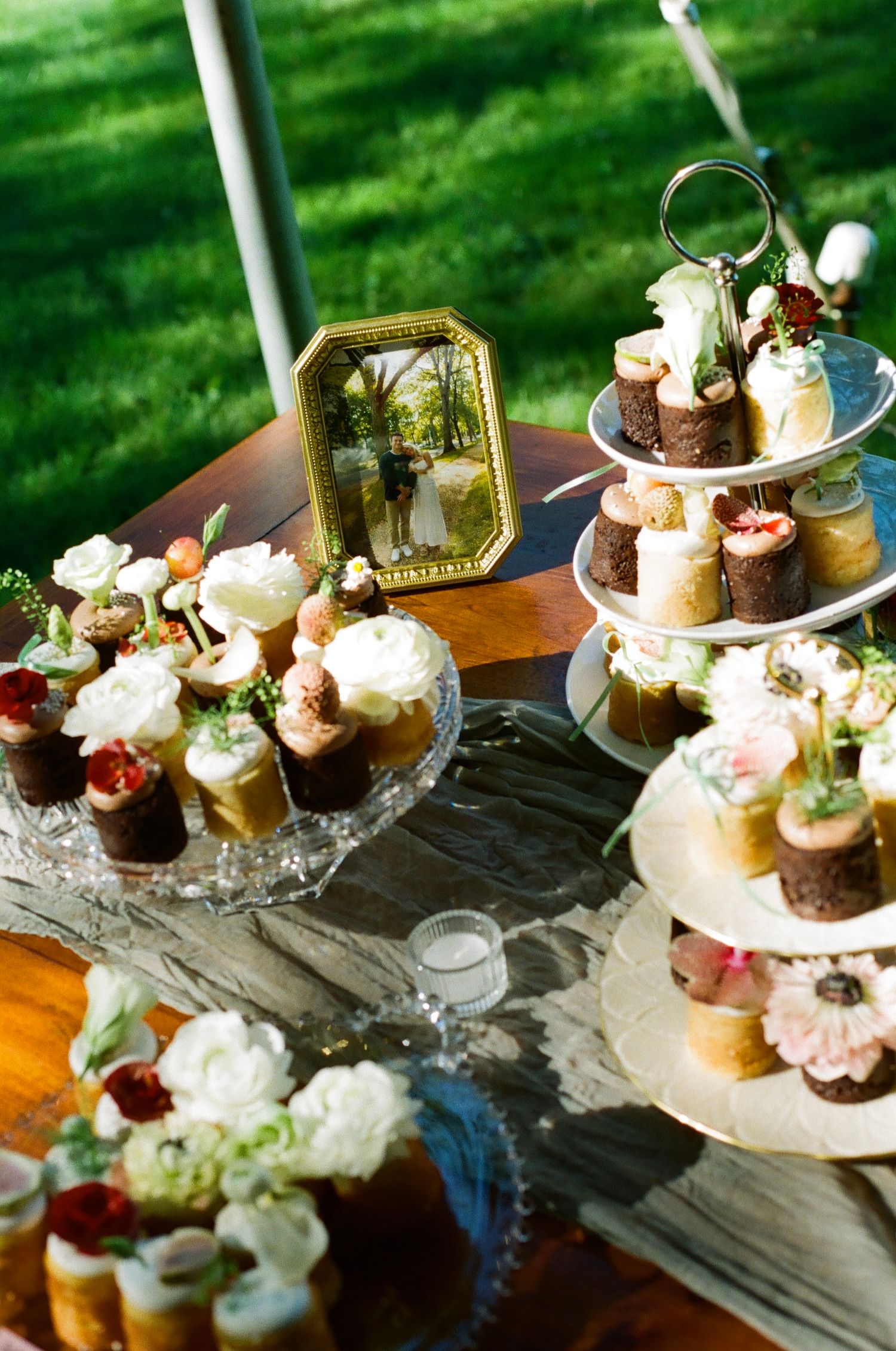 Elegant outdoor wedding dessert table with cupcakes, tiered stand, white flowers, and a framed couple photo on a wooden table.