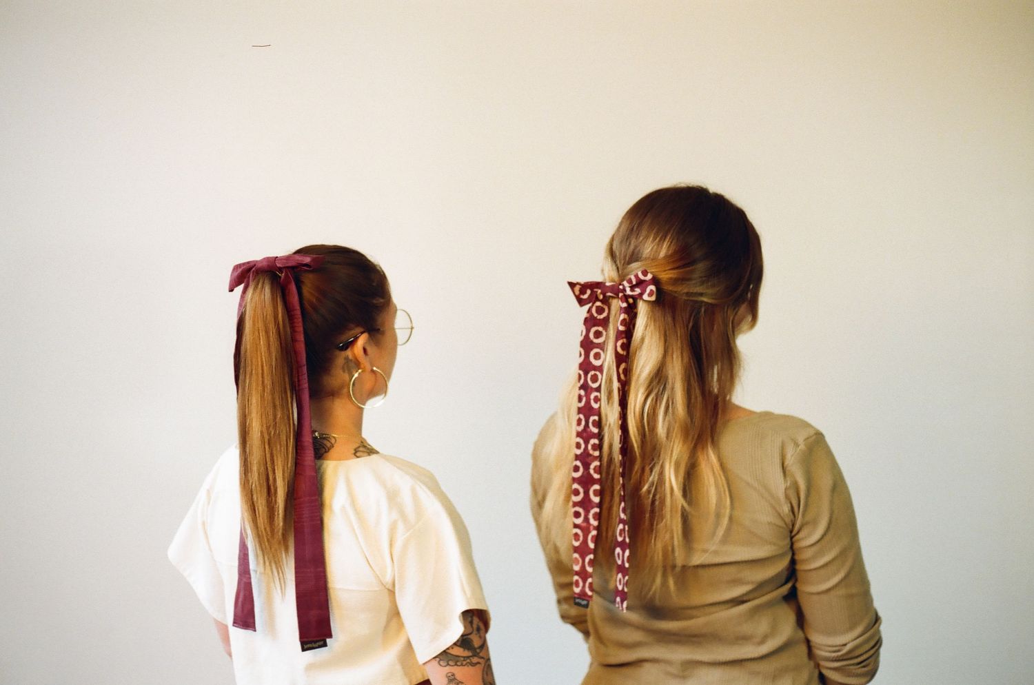 Two women seen from behind wearing burgundy hair bows and ribbons in their ponytails against a white wall.