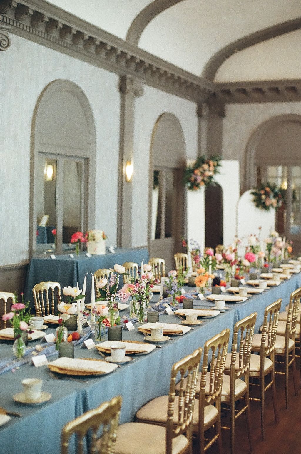 Elegant wedding reception with long blue-draped table, gold chiavari chairs, colorful floral centerpieces in a grand arched venue.