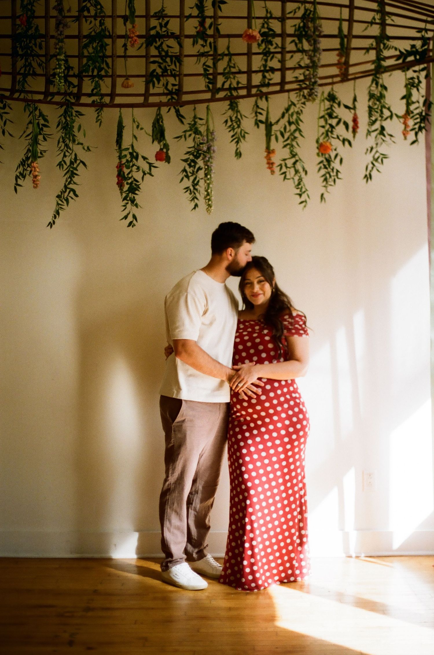 A pregnant woman in a red polka dot dress stands with her partner beneath hanging greenery in warm sunlight.