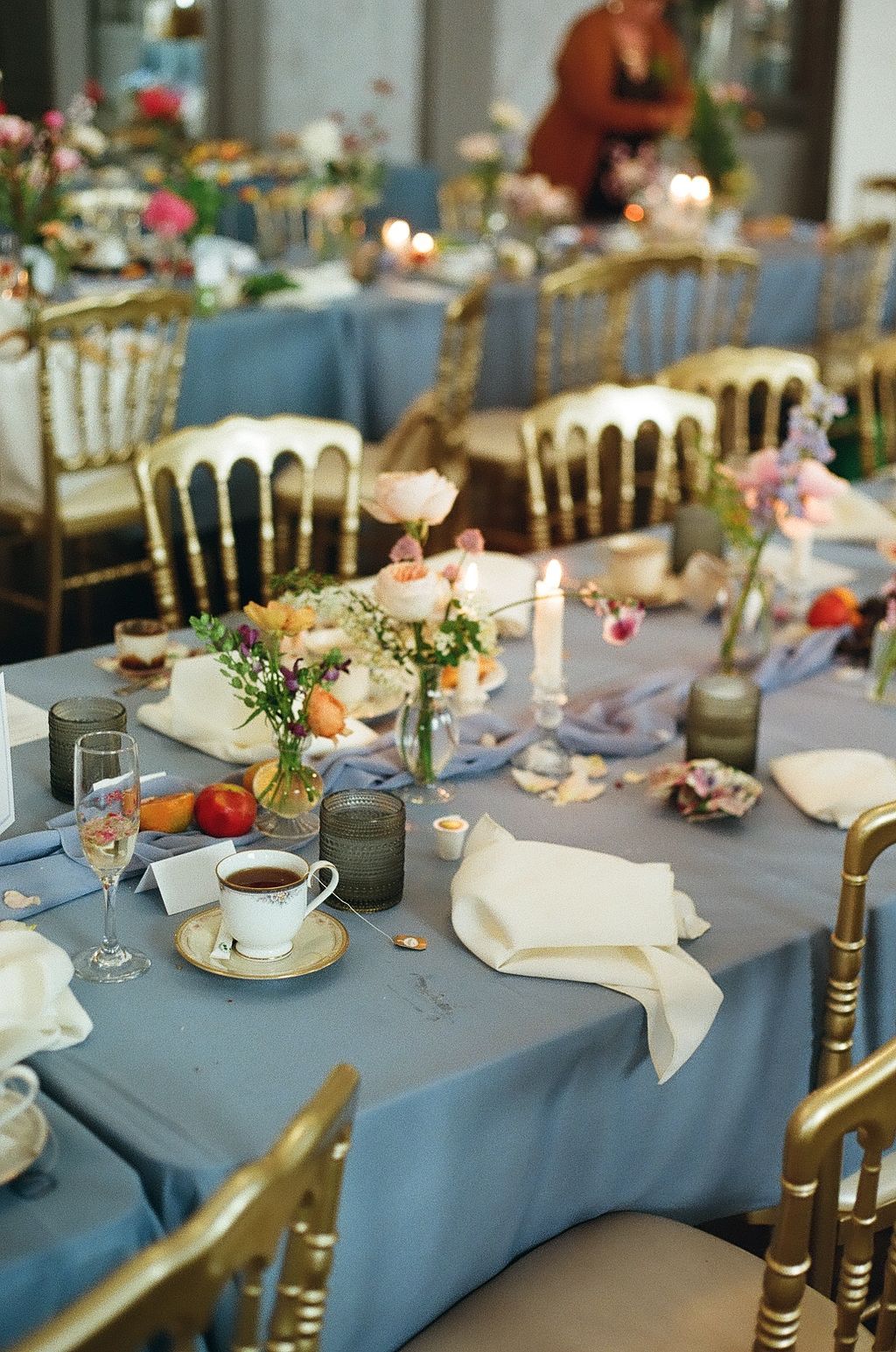 Elegant wedding reception table with dusty blue linens, gold chiavari chairs, floral centerpieces, candles, and white napkins.