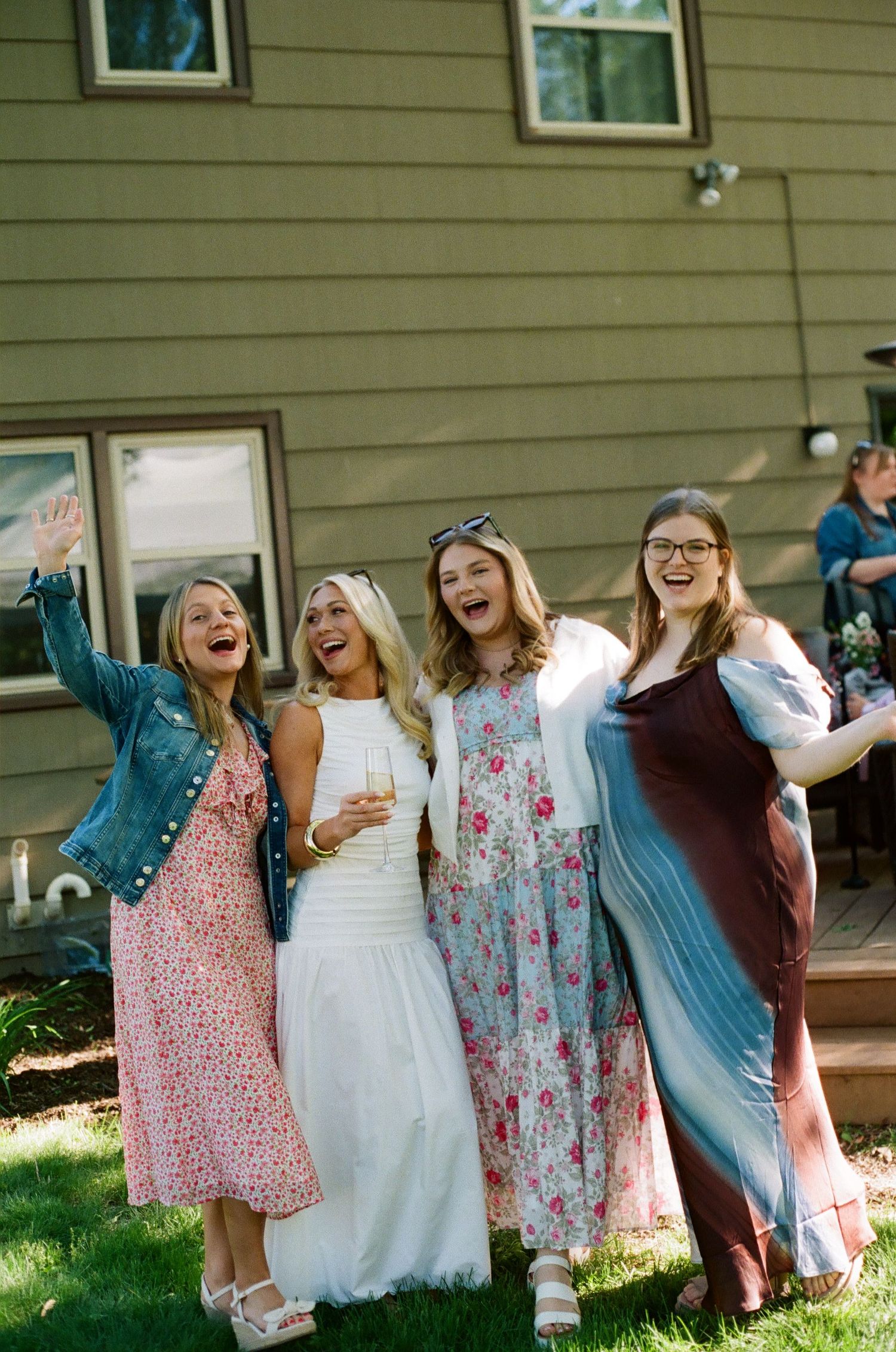 Four women in floral and colorful dresses laughing and posing together outdoors at a backyard celebration.