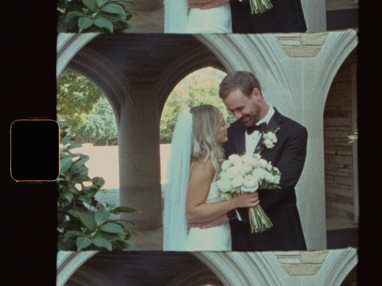 A bride and groom share a tender moment under stone arches, the bride holding a white bouquet in an elegant outdoor wedding setting.
