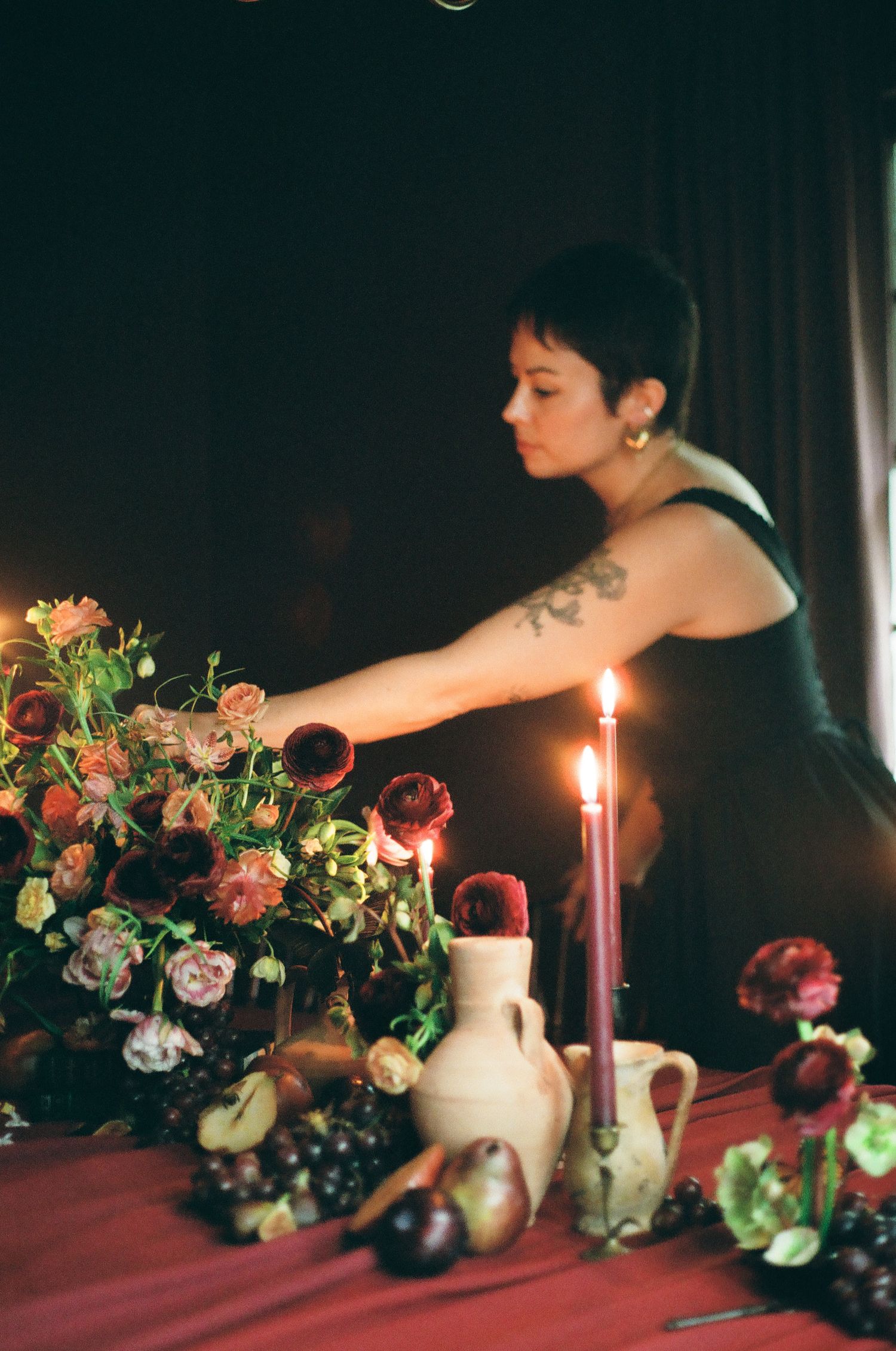 Woman arranging lush floral centerpiece with candles and fruit on a draped table in moody candlelit setting.