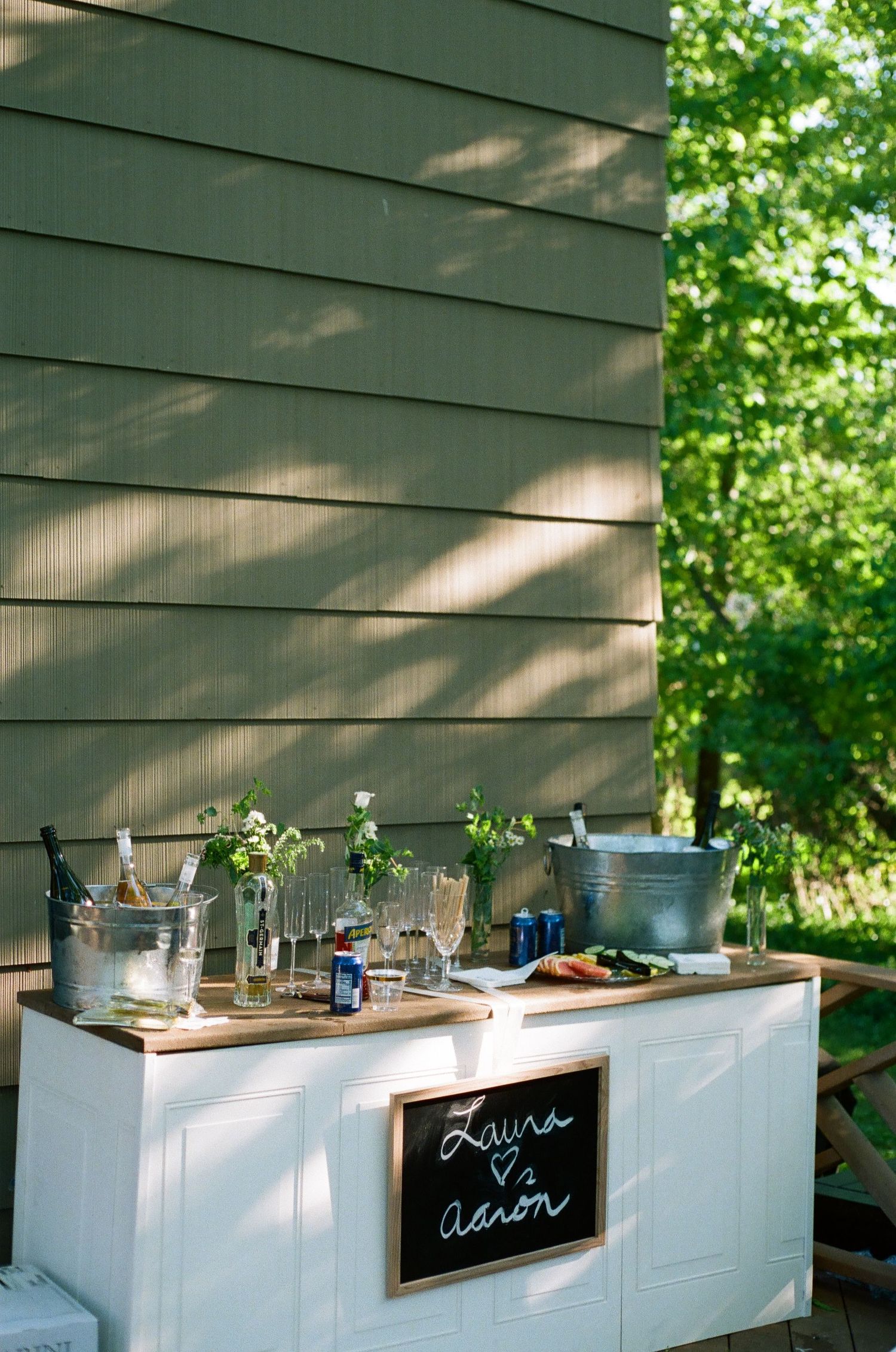 Outdoor wedding bar with chalkboard sign reading Laura & Aaron, galvanized buckets, glassware, and floral arrangements.