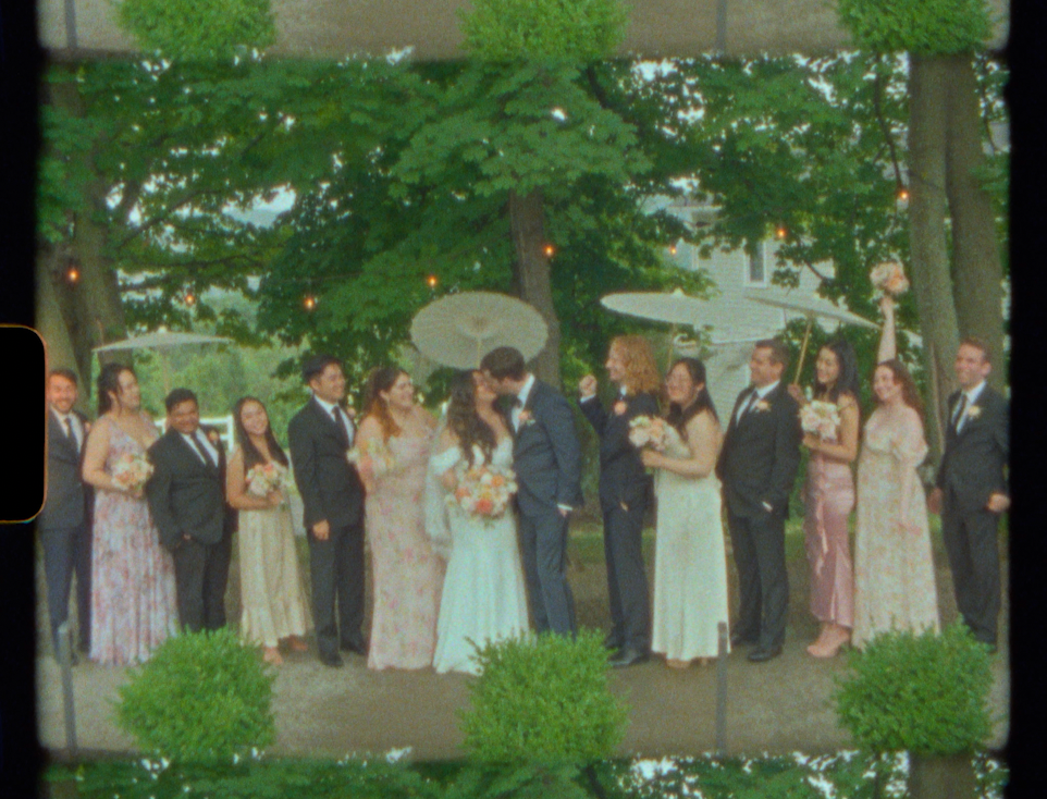 Wedding party group photo outdoors, bride and groom kissing surrounded by bridesmaids and groomsmen under trees.